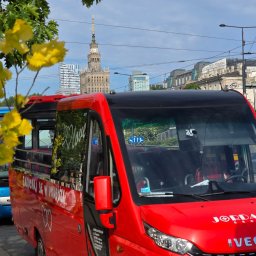 JACEK LEGENDZIEWICZ JORDAN GROUP - Czerwony autobus turystyczny z otwartym dachem na tle Pałacu Kultury w Warszawie, z napisem 'Zakochaj się w podróży' i logo firmy Jordan Group.