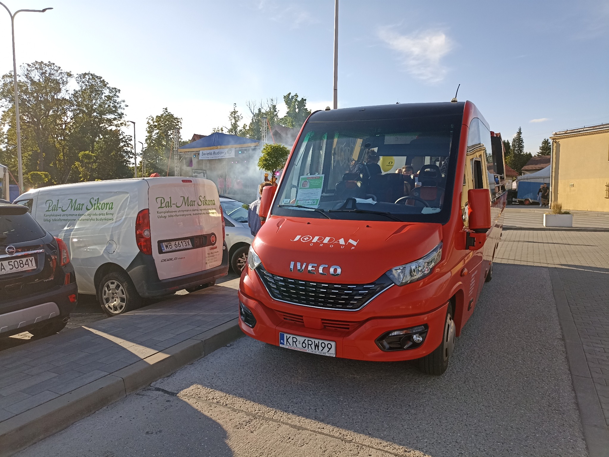 Czerwony bus Iveco Jordan Group z rejestracją KR 6RW99 na parkingu, obok biały furgon Pal-Mar Sikora. W tle scena z napisem Święto Rudawy i lekka mgła.