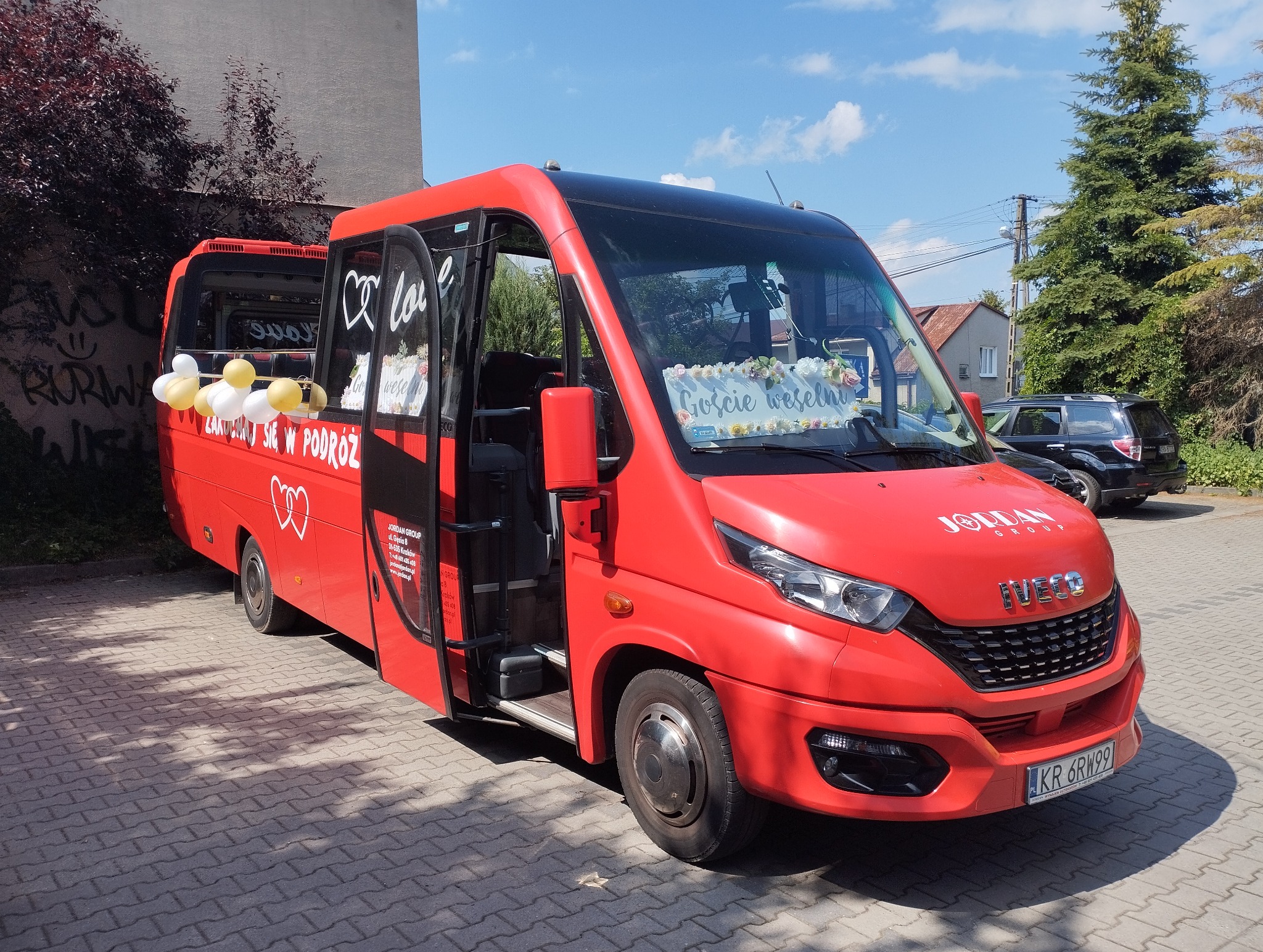Czerwony bus Iveco z dekoracją ślubną, otwarte drzwi, napis 'Goście weselni' na przedniej szybie, balony i napis 'Zakochani się w podróż'.