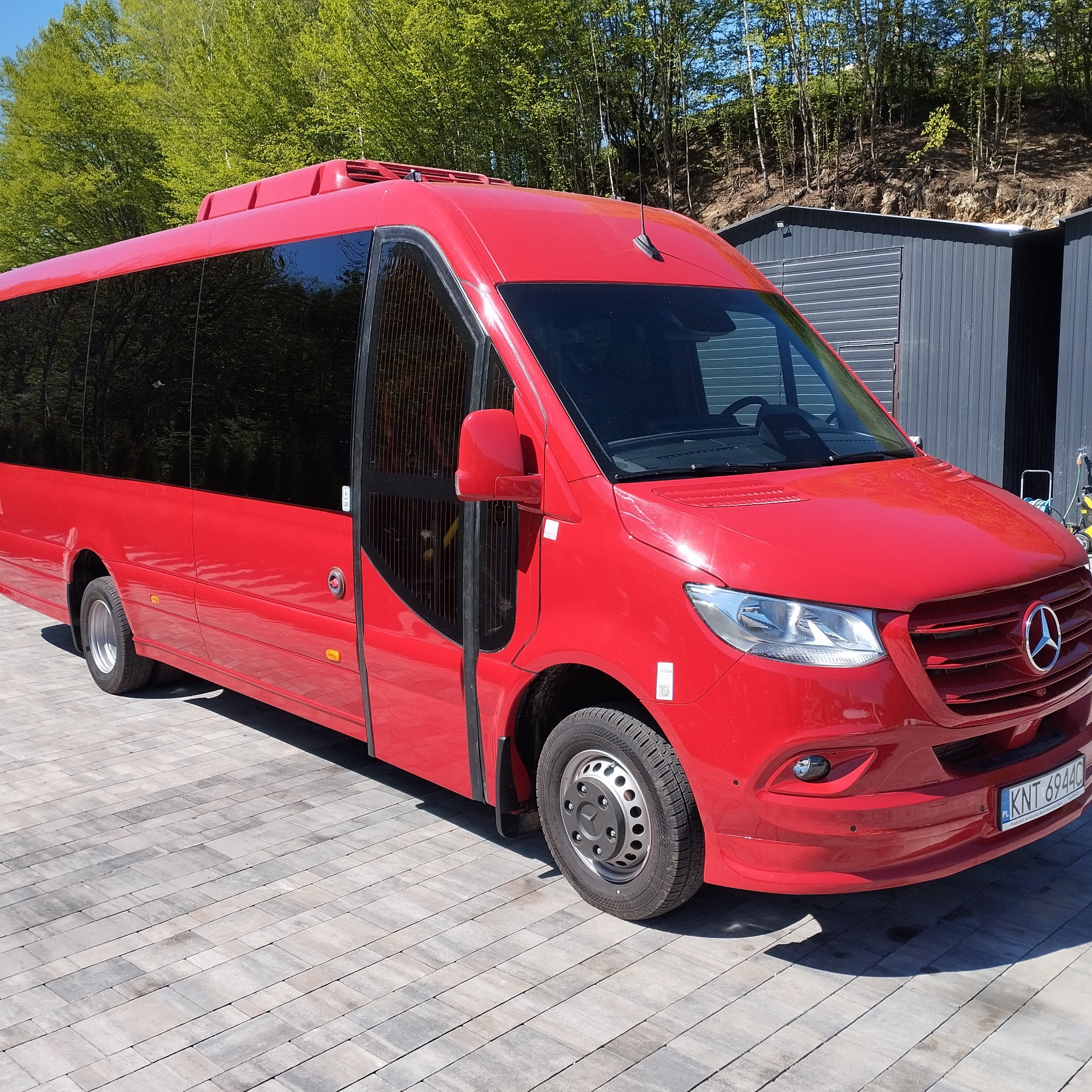 Czerwony bus Mercedes-Benz z otwartymi drzwiami, zaparkowany na zewnątrz, z zielonymi drzewami w tle. Widoczna tablica rejestracyjna.