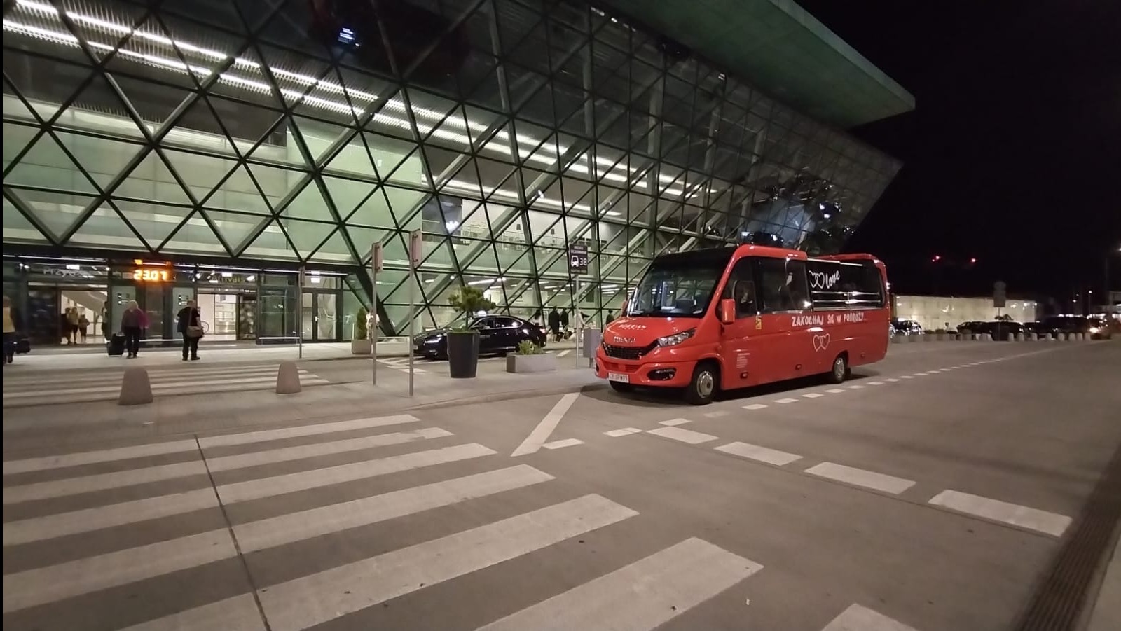 Czerwony bus z napisem 'Zakochaj się w podróży' stoi przed nowoczesnym budynkiem lotniska nocą. Widoczne przejście dla pieszych i sylwetki podróżnych.