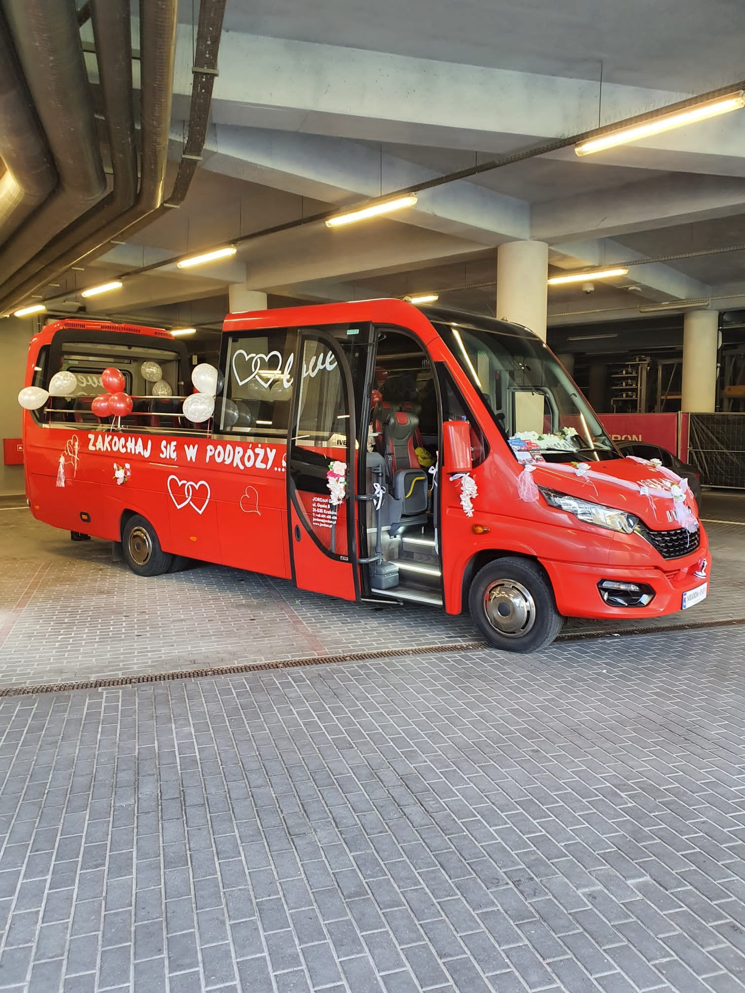 Czerwony, udekorowany bus z otwartymi drzwiami, z napisem 'Zakochaj się w podróży...' i balonami. Dekoracja ślubna, przewóz osób.