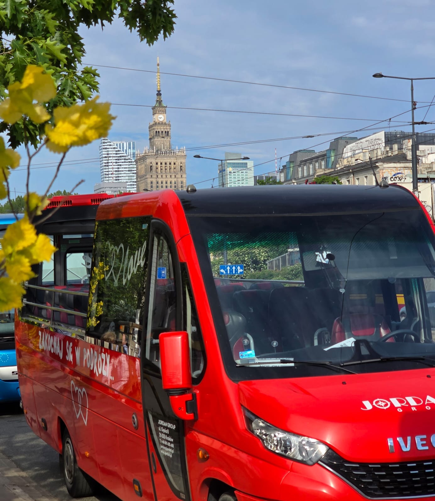 Czerwony autobus turystyczny z otwartym dachem na tle Pałacu Kultury w Warszawie, z napisem 'Zakochaj się w podróży' i logo firmy Jordan Group.