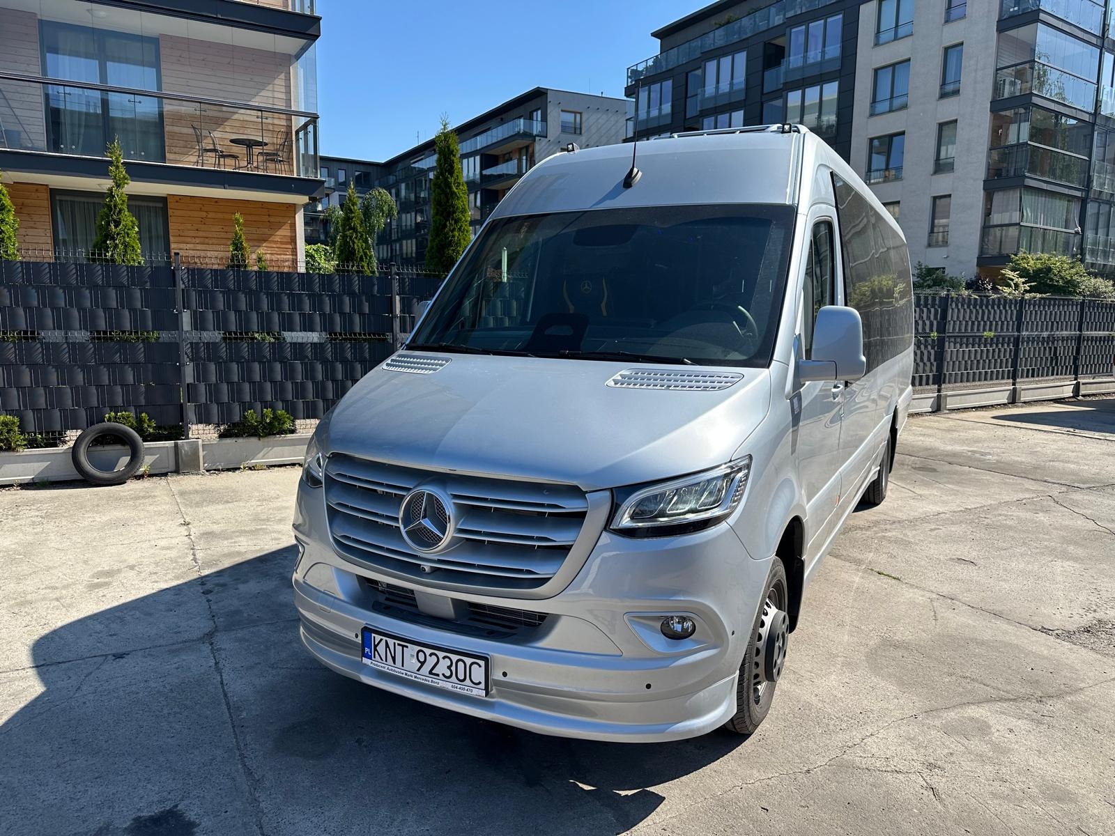Srebrny Mercedes-Benz Sprinter z anteną na dachu, zaparkowany na tle nowoczesnych budynków. Widoczna tablica rejestracyjna i opona oparta o płot.