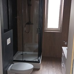 IVAN ANDRUSYSHYN - Nowoczesna łazienka po remoncie: kabina prysznicowa z czarną armaturą, toaleta podwieszana, drewnopodobne płytki podłogowe i okno z białymi ramami.