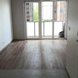 IVAN ANDRUSYSHYN - Jasne wnętrze z drewnopodobną podłogą i szarą płytką, widok na przeszklony balkon. W tle budynki.