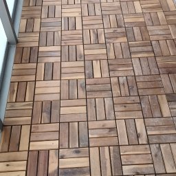 IVAN ANDRUSYSHYN - Ułożone drewniane płytki podłogowe na balkonie, widok z góry, zróżnicowany odcień drewna, regularny wzór, szara balustrada po lewej stronie.