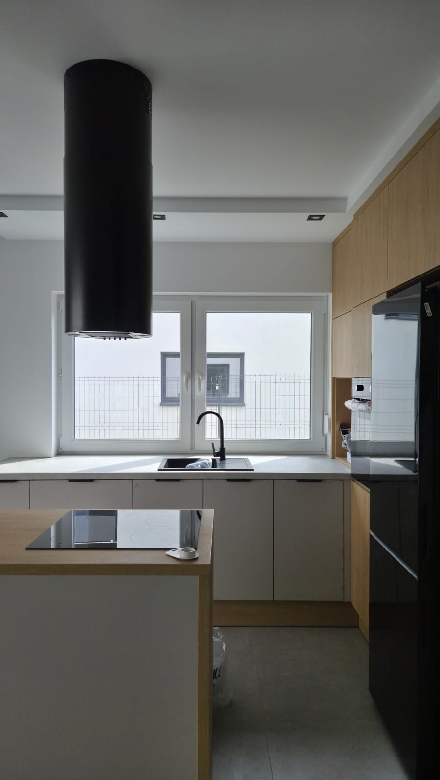Nowoczesna kuchnia z wyspą i czarnym okapem cylindrycznym. Jasne szafki, biały blat, okno z widokiem na ogrodzenie. Minimalistyczny design.