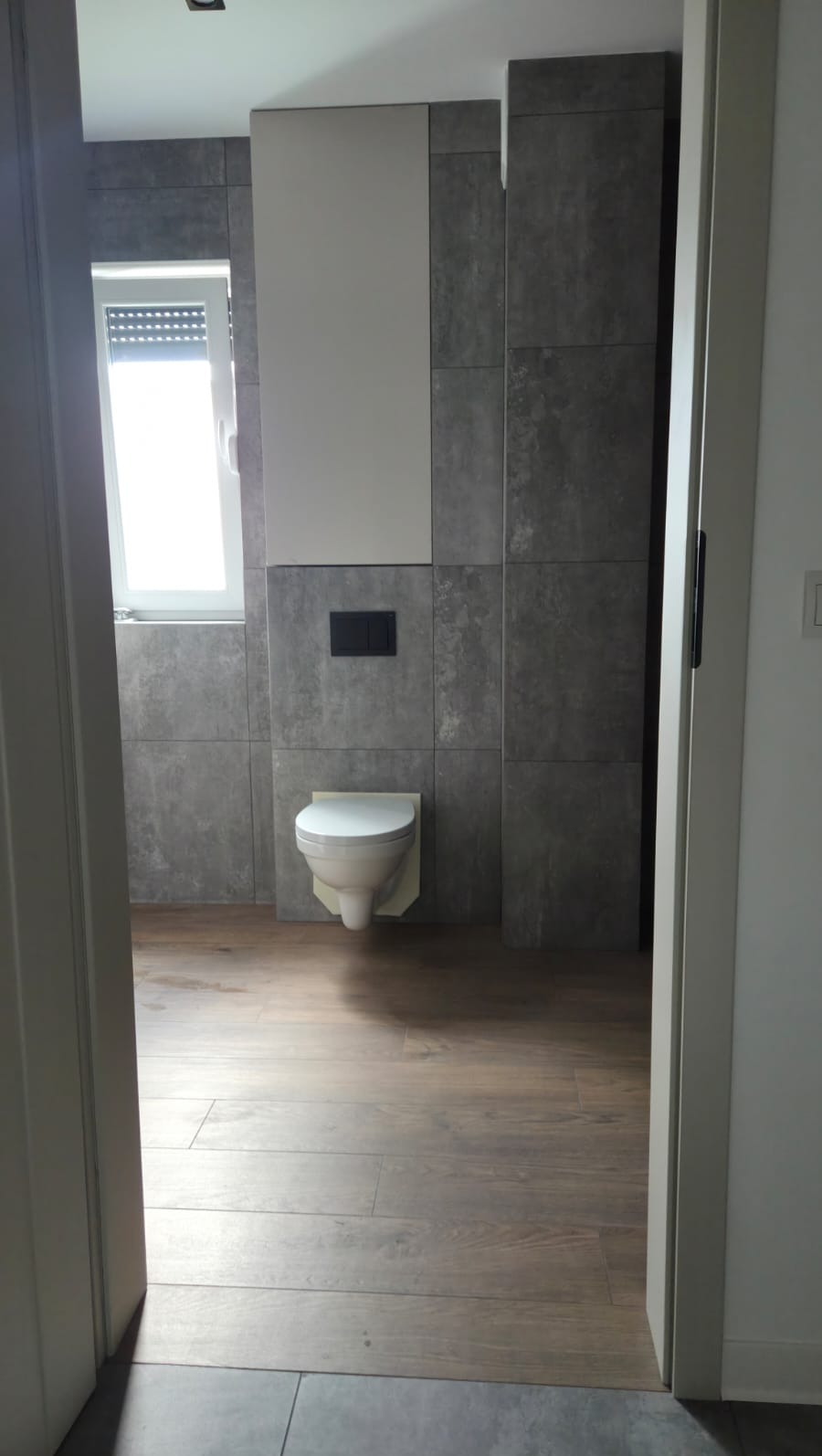 Nowoczesna łazienka z drewnopodobną podłogą i szarą płytką na ścianie. Wisząca toaleta, okno z roletami. Widok zza otwartych drzwi.