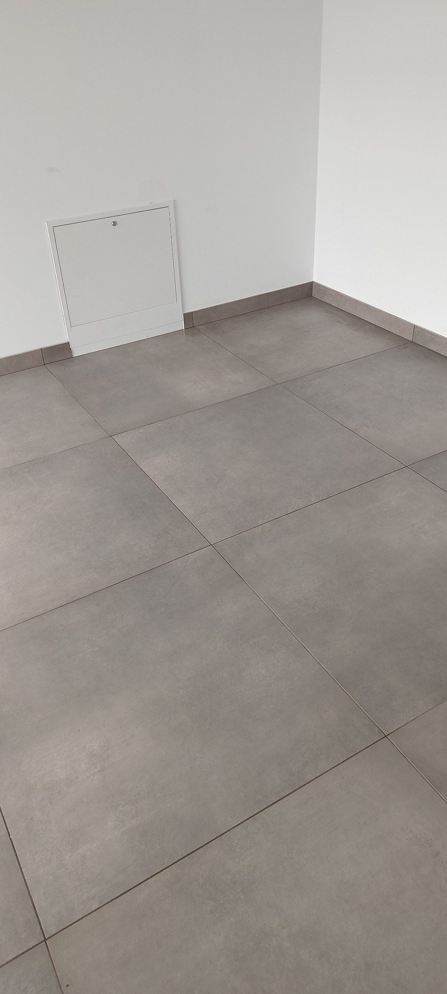 Szare, matowe płytki podłogowe ułożone w minimalistycznym wnętrzu. Widoczna biała ściana z dostępem rewizyjnym i szare listwy przypodłogowe.