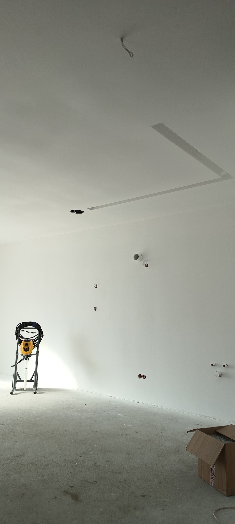 Wnętrze w trakcie prac wykończeniowych: białe ściany, surowa podłoga, otwory na instalacje i sprzęt do malowania natryskowego. Widoczny fragment podwieszanego sufitu.