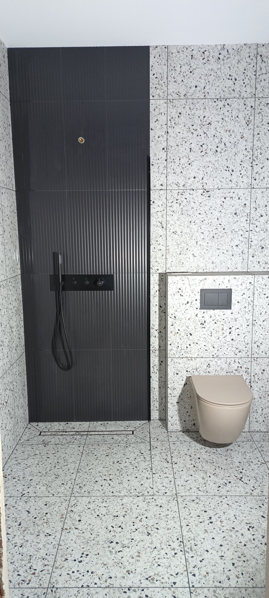Nowoczesna łazienka: czarna armatura prysznicowa na tle pionowych, grafitowych płytek, obok toaleta i ściany wykończone płytkami lastryko.