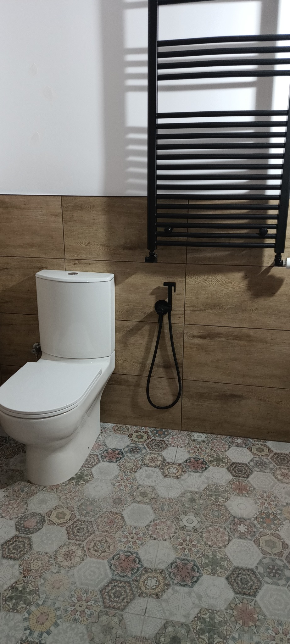 Wykończona toaleta z płytkami imitującymi drewno i wzorzystą podłogą. Widoczny biały sedes i czarny grzejnik drabinkowy z bidetką. Nowoczesny design.