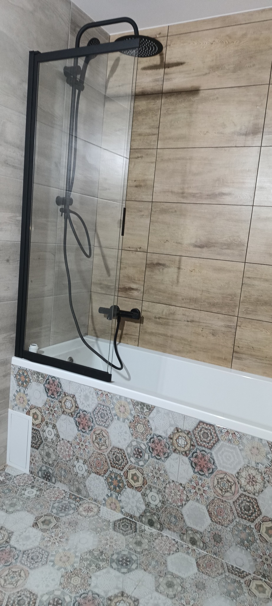 Nowoczesna kabina prysznicowa z czarną armaturą i dekoracyjnymi płytkami heksagonalnymi na obudowie wanny i podłodze, drewnopodobne płytki na ścianach.