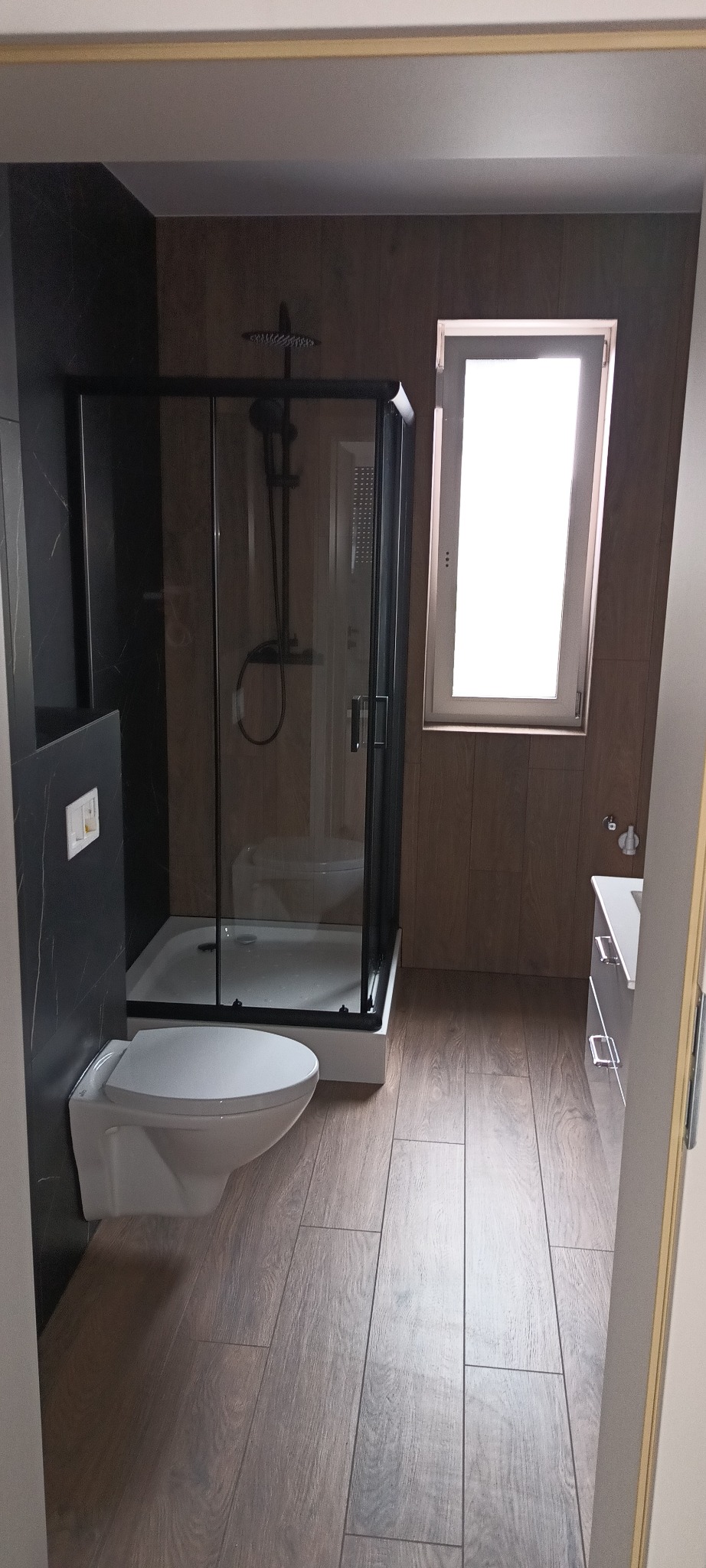 Nowoczesna łazienka po remoncie: kabina prysznicowa z czarną armaturą, toaleta podwieszana, drewnopodobne płytki podłogowe i okno z białymi ramami.