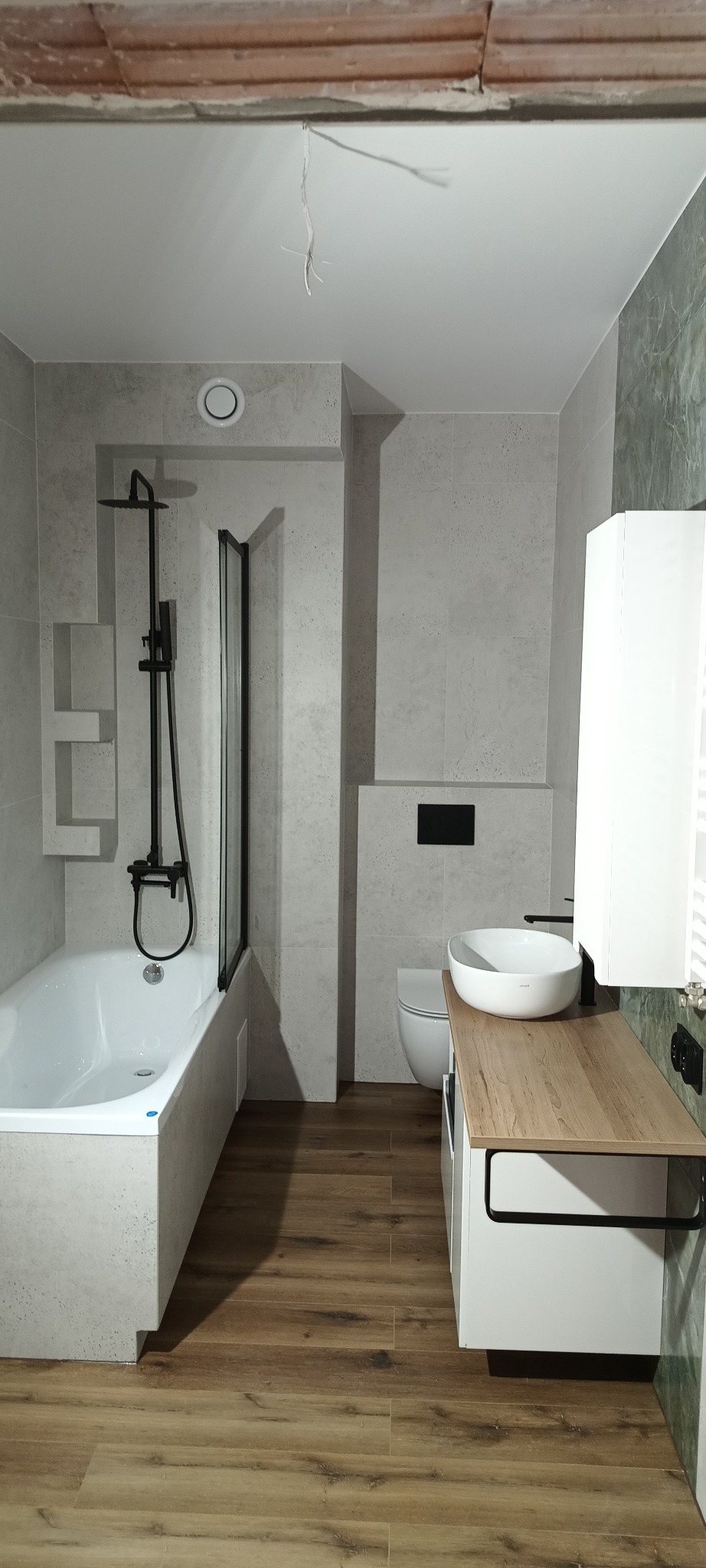 Nowoczesna łazienka z drewnianą podłogą i jasnymi płytkami. Wanna z czarną armaturą i szklanym parawanem. Umywalka nablatowa na szafce z blatem.