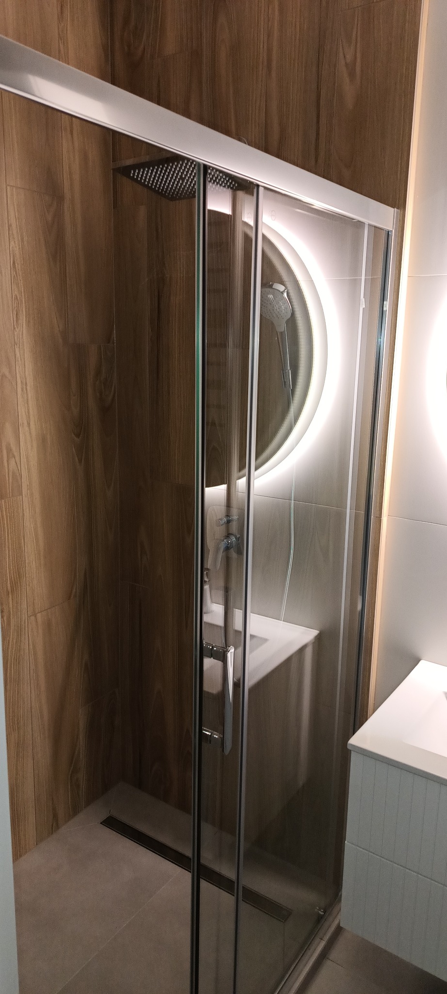 Nowoczesna kabina prysznicowa ze szklanymi drzwiami przesuwnymi, deszczownicą i okrągłym lustrem z podświetleniem LED. Ściany wyłożone płytkami imitującymi drewno.