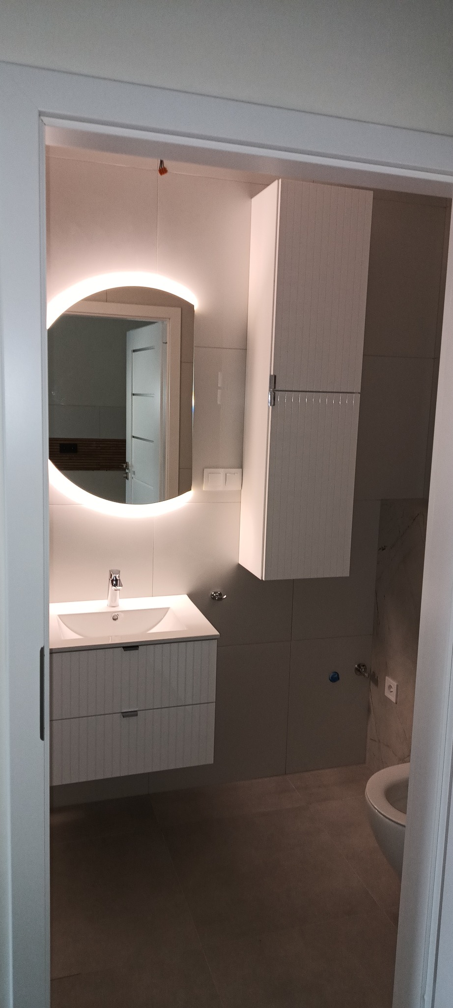 Nowoczesna łazienka z podświetlanym lustrem, umywalką z szafką, wiszącą szafką i szarą glazurą. Widok przez otwarte drzwi.