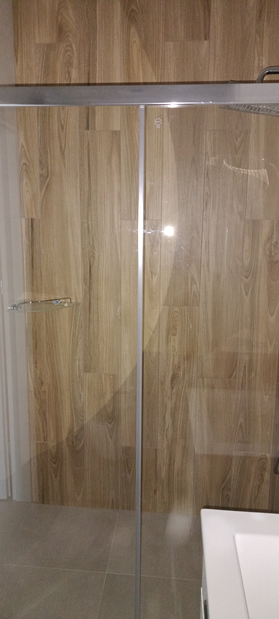 Kabina prysznicowa z drewnopodobnymi płytkami na ścianach, widoczne srebrne profile i szklane drzwi. Umywalka w rogu kadru.