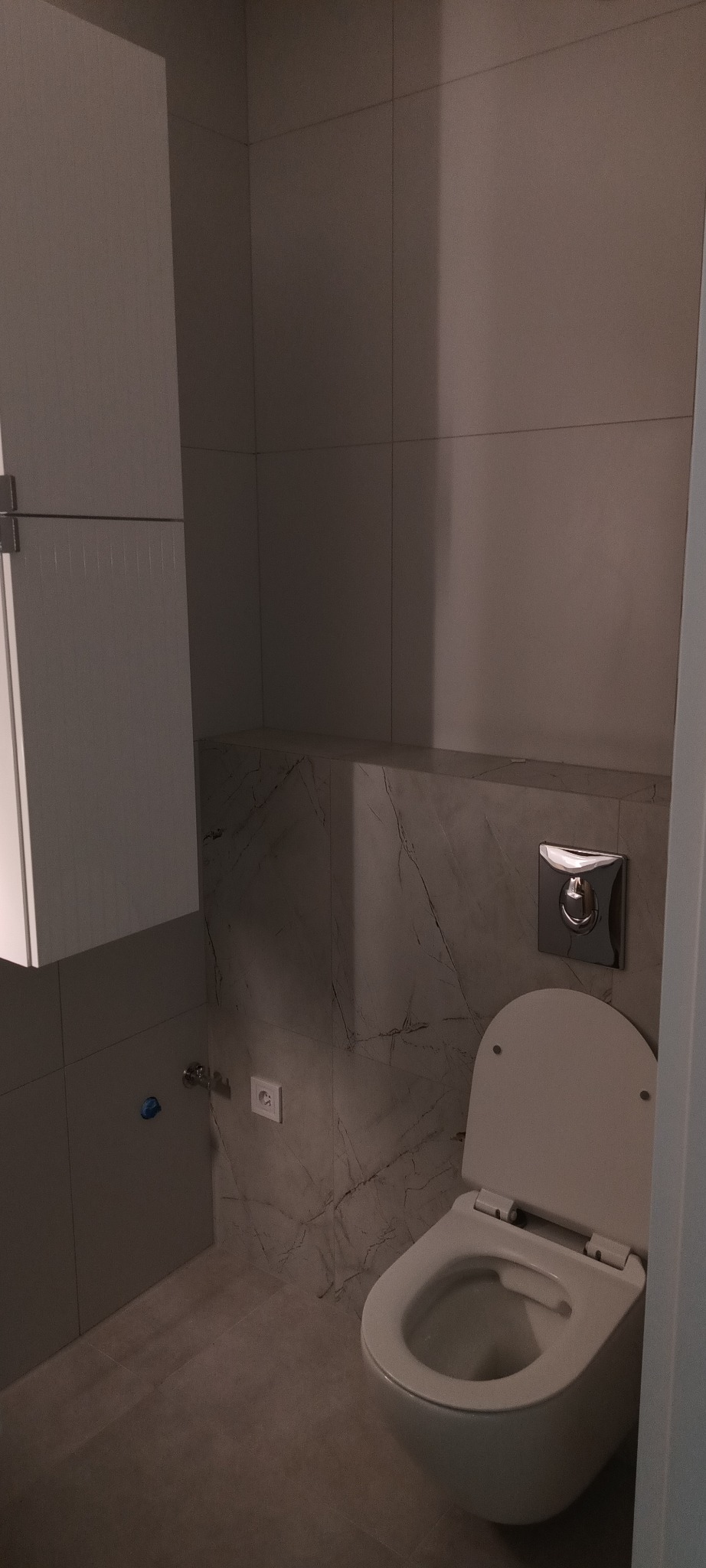 Nowoczesna toaleta wisząca z zabudową i marmurowymi płytkami. Widoczny przycisk spłukiwania i gniazdko elektryczne. Minimalistyczny design łazienki.