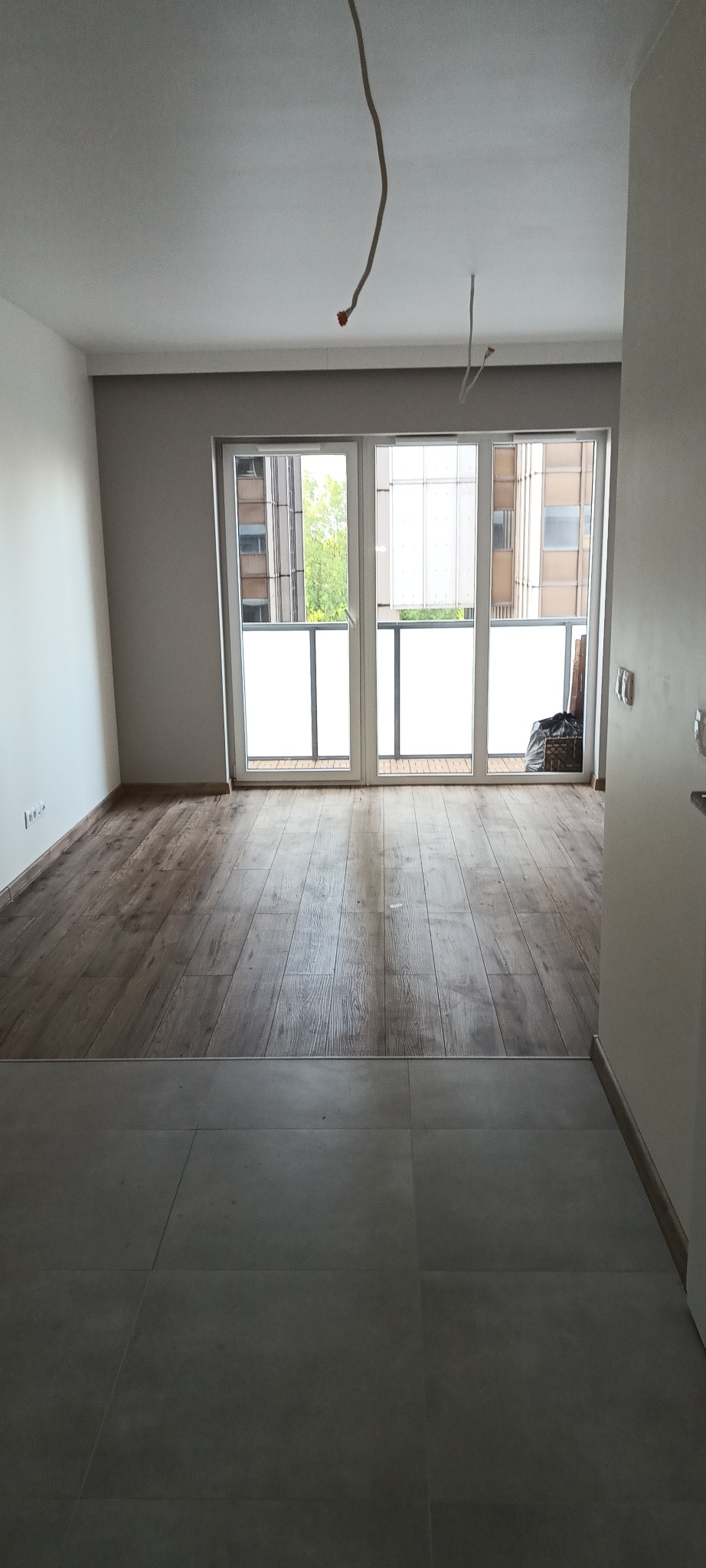 Jasne wnętrze z drewnopodobną podłogą i szarą płytką, widok na przeszklony balkon. W tle budynki.