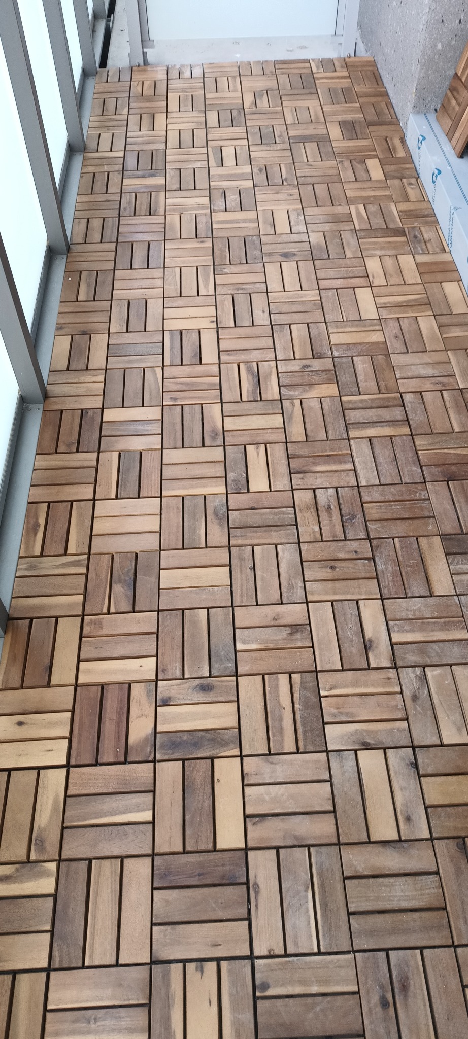 Ułożone drewniane płytki podłogowe na balkonie, widok z góry, zróżnicowany odcień drewna, regularny wzór, szara balustrada po lewej stronie.