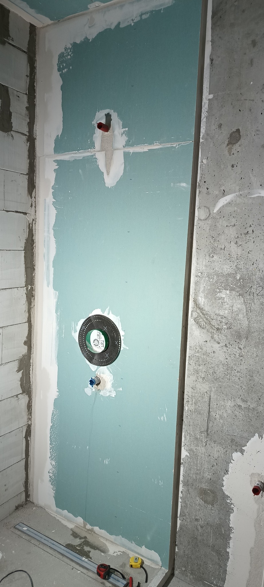 Niebieska płyta G-K z instalacją wodną w surowej łazience. Widoczne narzędzia pomiarowe i ślady szpachlowania. Ściana z betonu komórkowego i surowy beton.