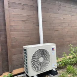 Zamontowana jednostka zewnętrzna klimatyzacji Gree na betonowych podstawach przy drewnianej ścianie budynku z rurą odprowadzającą. Widoczne rośliny w tle.