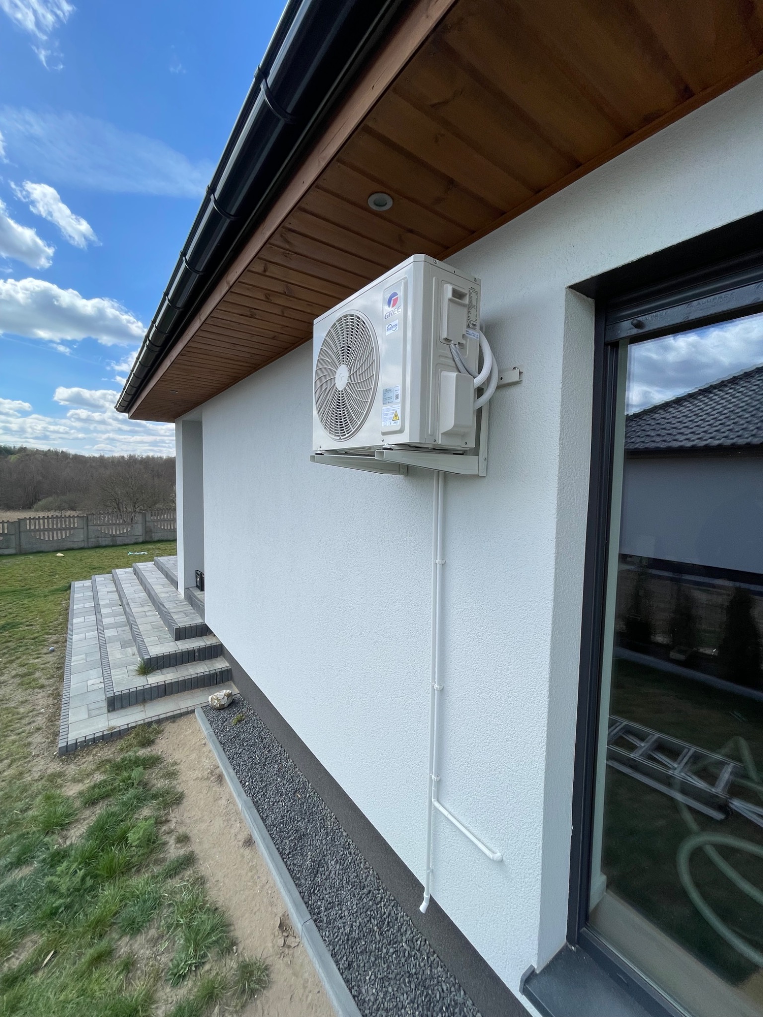Biała jednostka zewnętrzna klimatyzacji Gree zamontowana na elewacji domu z widocznymi przewodami. W tle fragment schodów wejściowych i okno.