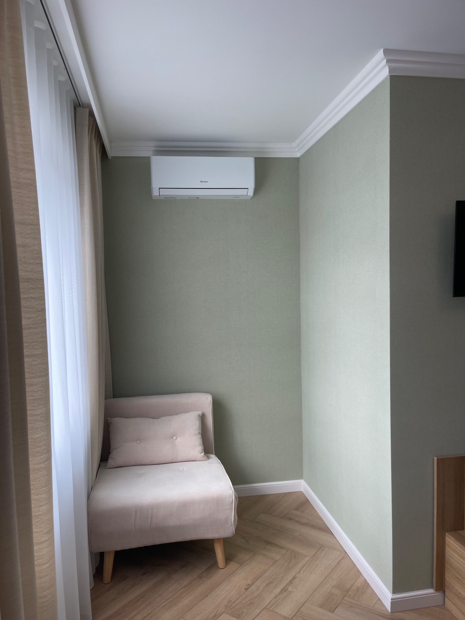 Wnętrze pokoju z klimatyzatorem ściennym, beżowym fotelem i zasłonami. Parkiet ułożony we wzór jodełki. Ściany w odcieniach zieleni, białe listwy przypodłogowe i sufitowe.