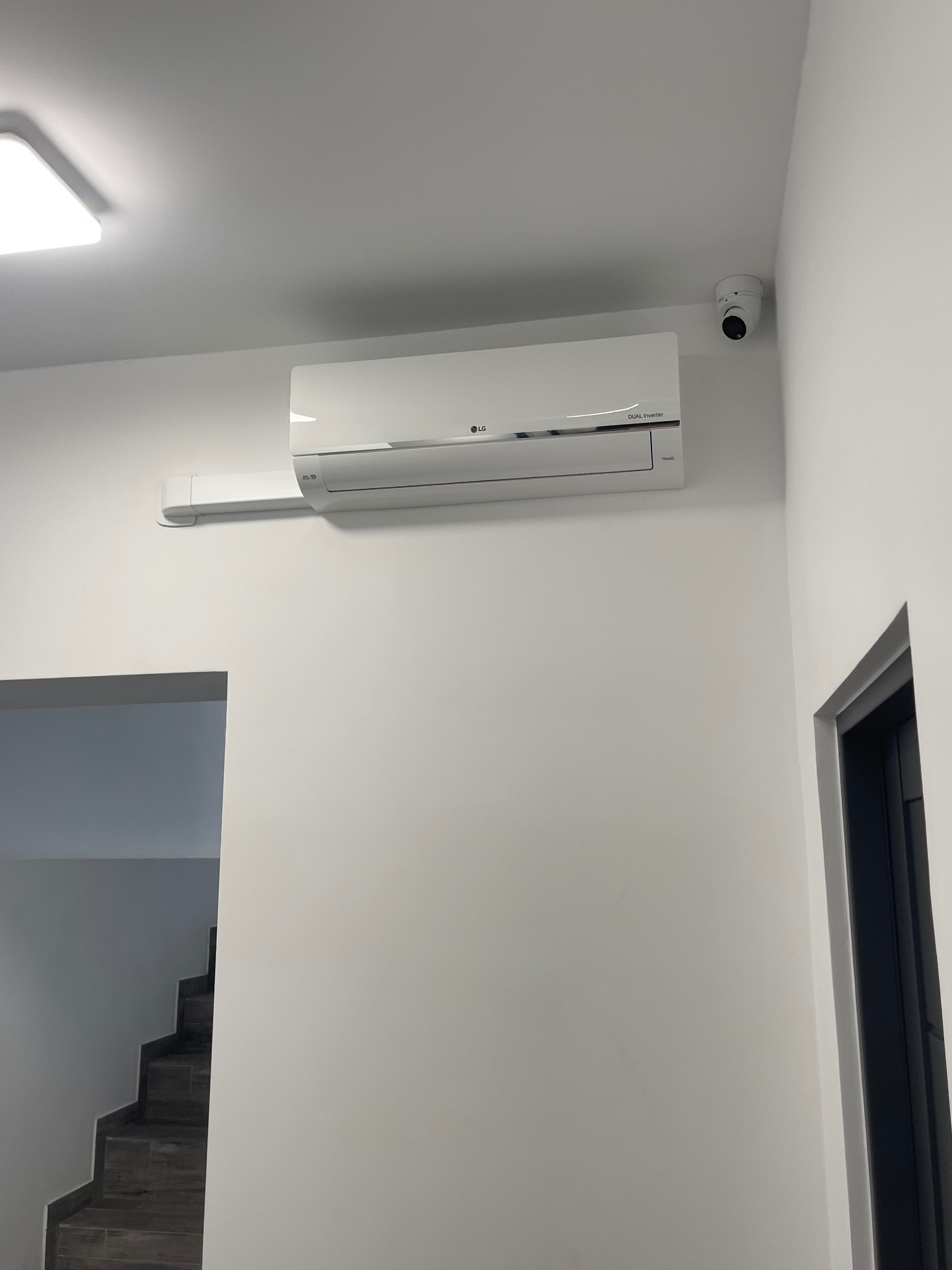 Biała klimatyzacja LG Dual Inverter zamontowana na ścianie, widoczna kamera monitoringu i fragment schodów. Minimalistyczne wnętrze.