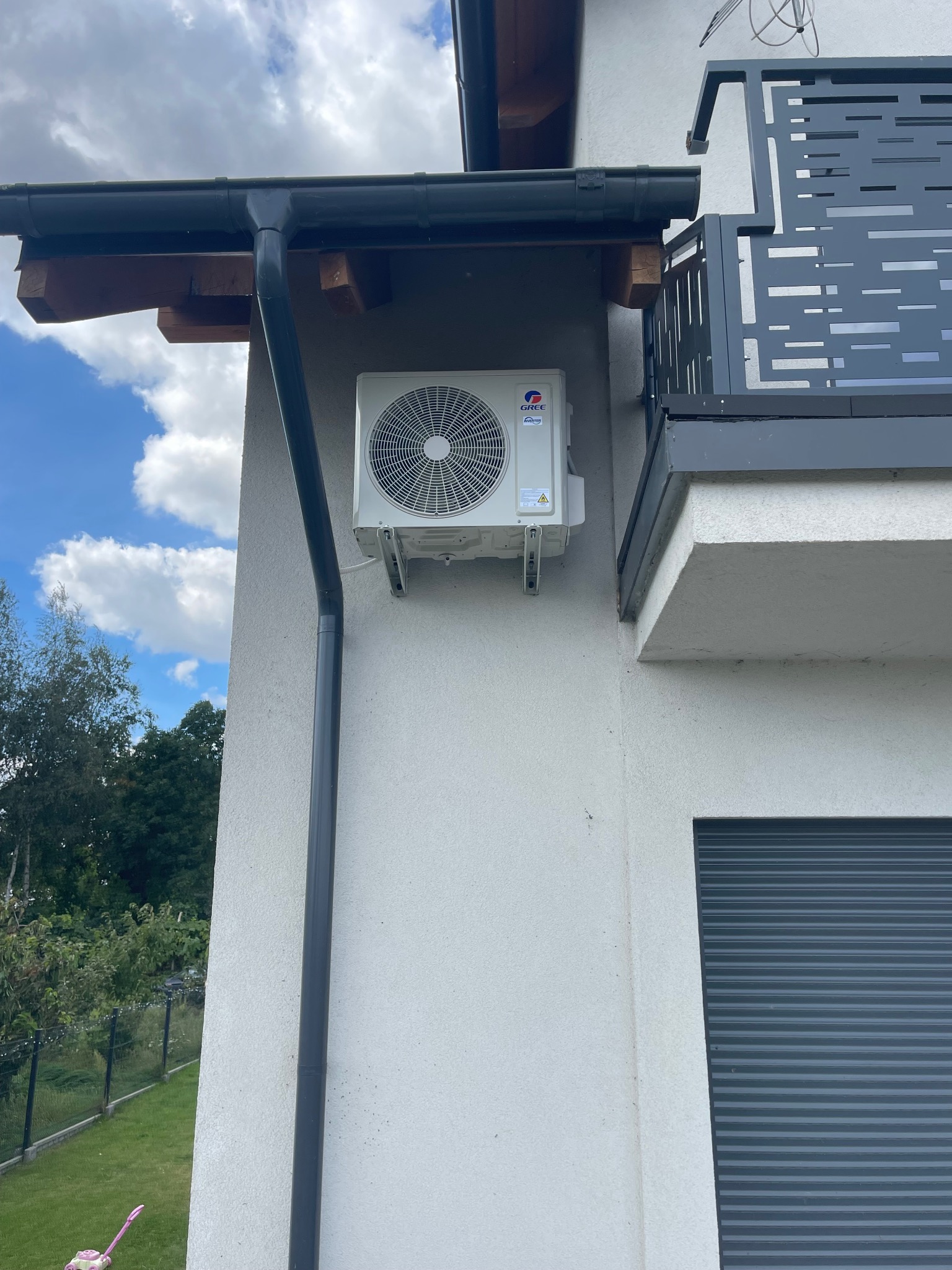 Biała klimatyzacja Gree zamontowana na elewacji domu z szarą rynną i balkonem. Widoczne mocowania i fragment okna z roletą. Błękitne niebo w tle.