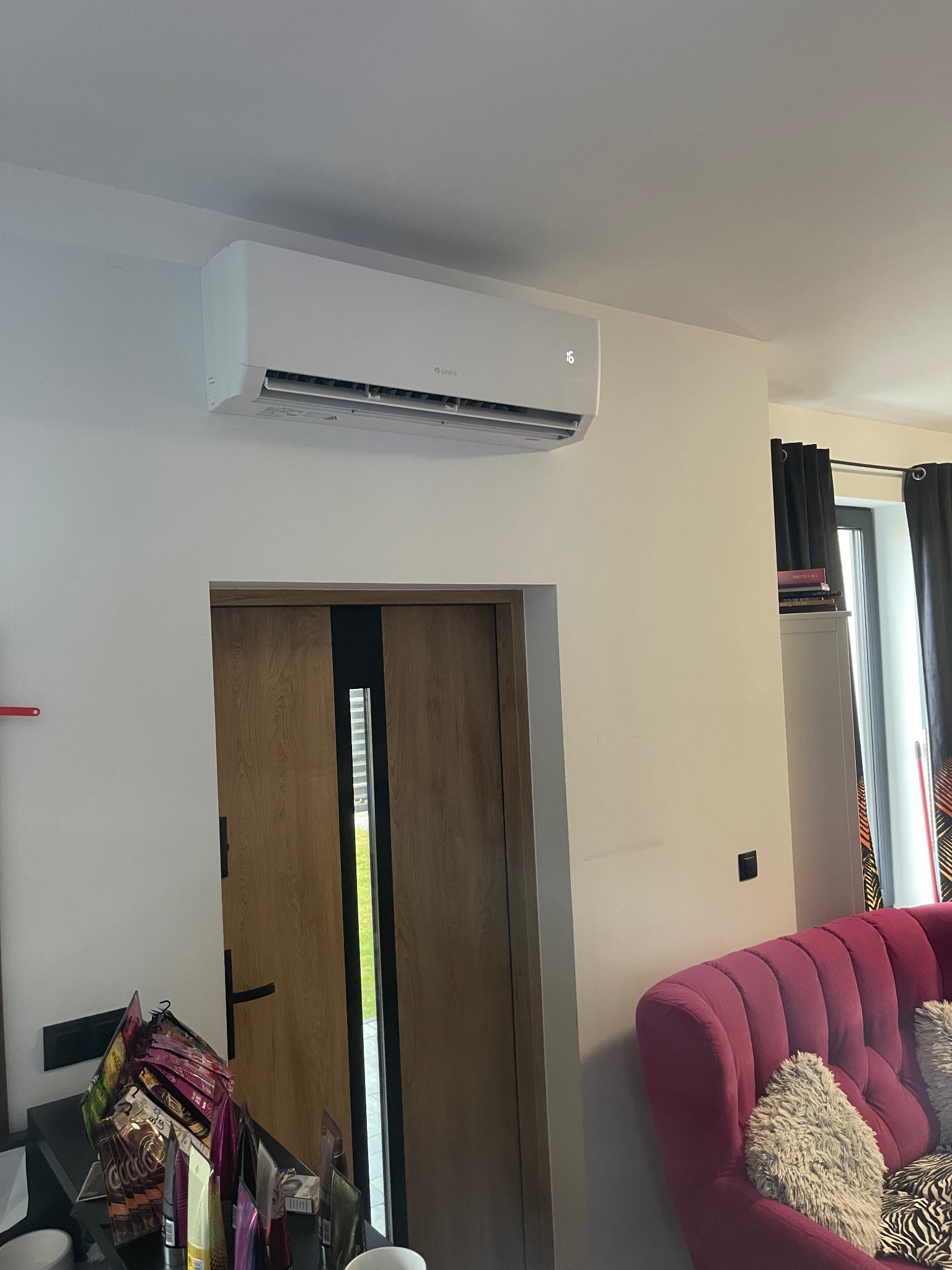 Biała klimatyzacja ścienna z wyświetlaczem temperatury (16 stopni) zamontowana nad drzwiami w jasnym wnętrzu. Widoczny fragment różowego fotela i okna.