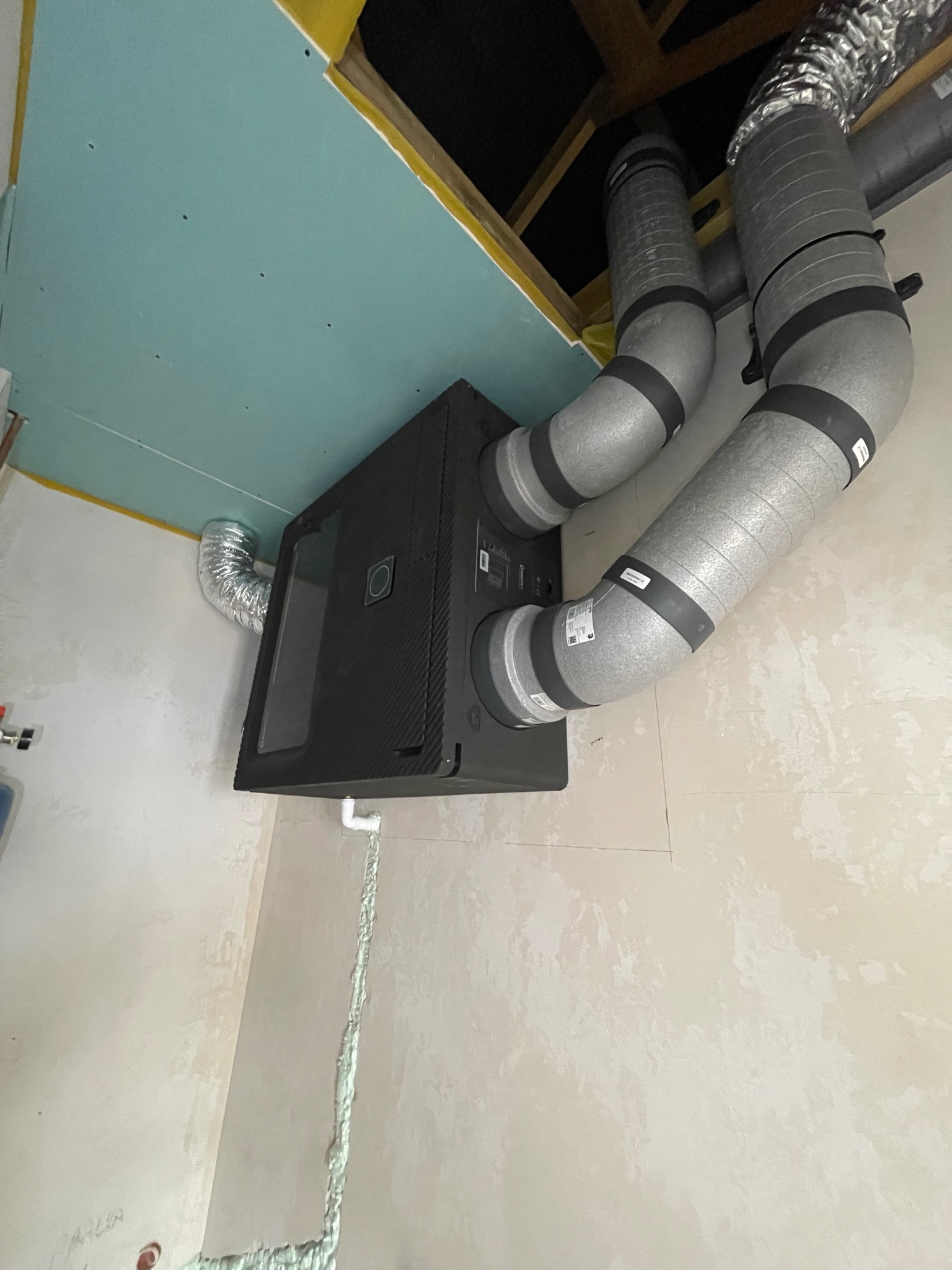 Czarny rekuperator zamontowany na ścianie z podłączonymi izolowanymi przewodami wentylacyjnymi, widok z dołu, sufit z płyt karton-gips.