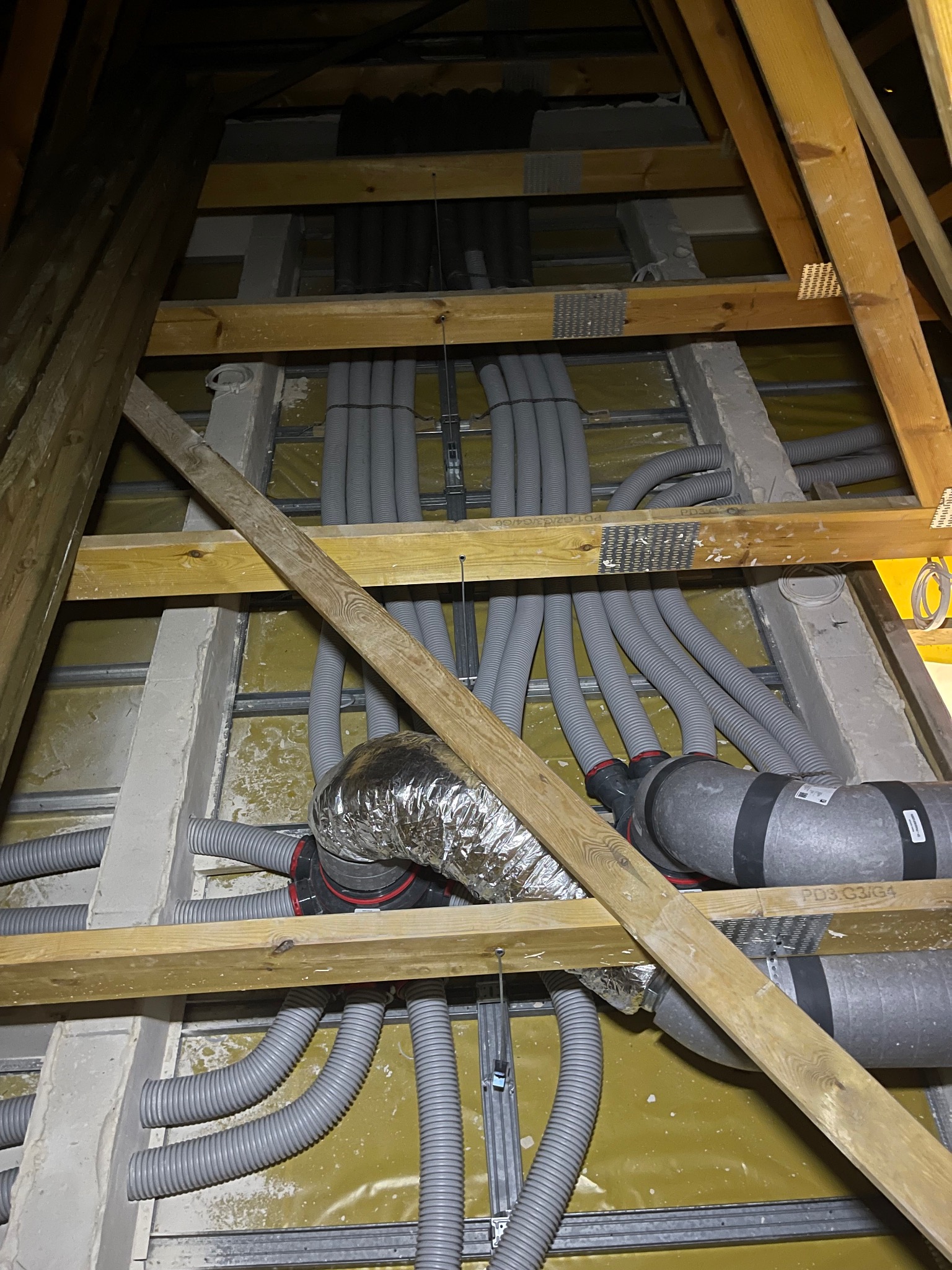 Szare, elastyczne rury wentylacyjne rozprowadzone w przestrzeni poddasza, widoczne elementy konstrukcji drewnianej i metalowej. Instalacja w trakcie montażu.