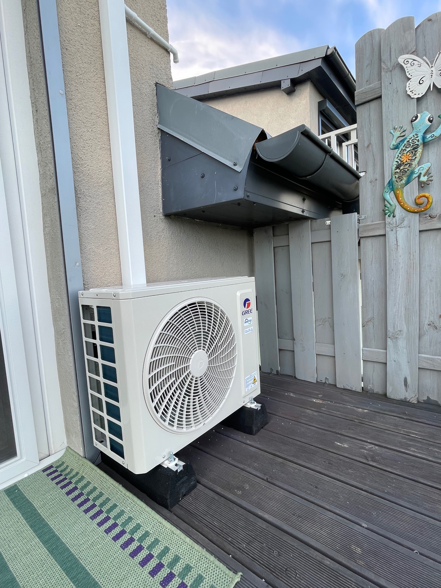 Biały klimatyzator GREE zamontowany na zewnątrz budynku, na drewnianym tarasie, z widocznymi elementami instalacji i dekoracyjną ozdobą na płocie.