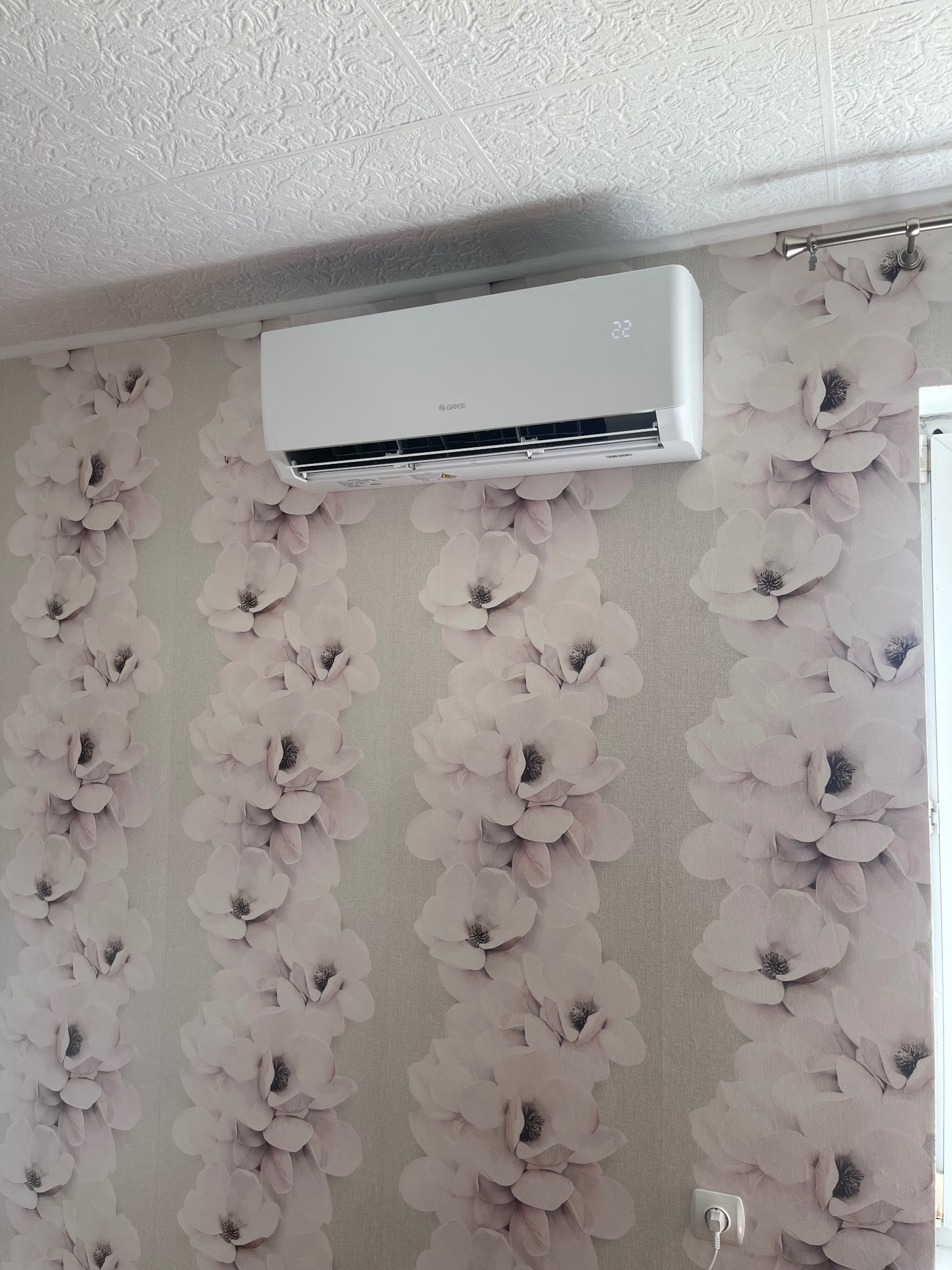 Biała klimatyzacja Gree na ścianie z tapetą w kwiaty magnolii. Widoczny fragment sufitu i karnisza. Nowoczesny design, temperatura ustawiona na 22 stopnie.