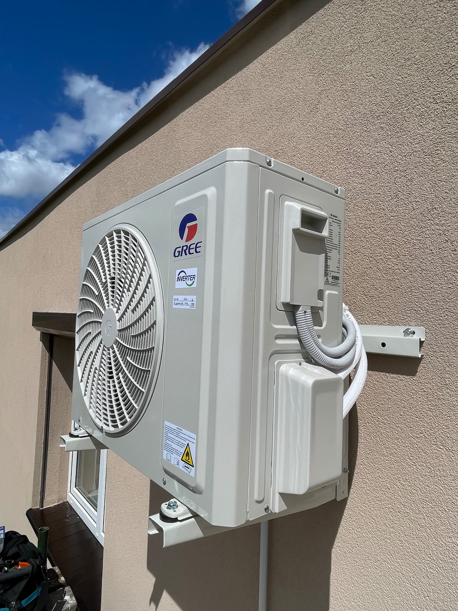 Zamontowana jednostka zewnętrzna klimatyzacji Gree na beżowej elewacji budynku, widoczne okno i fragment narzędzi. Błękitne niebo z chmurami w tle.