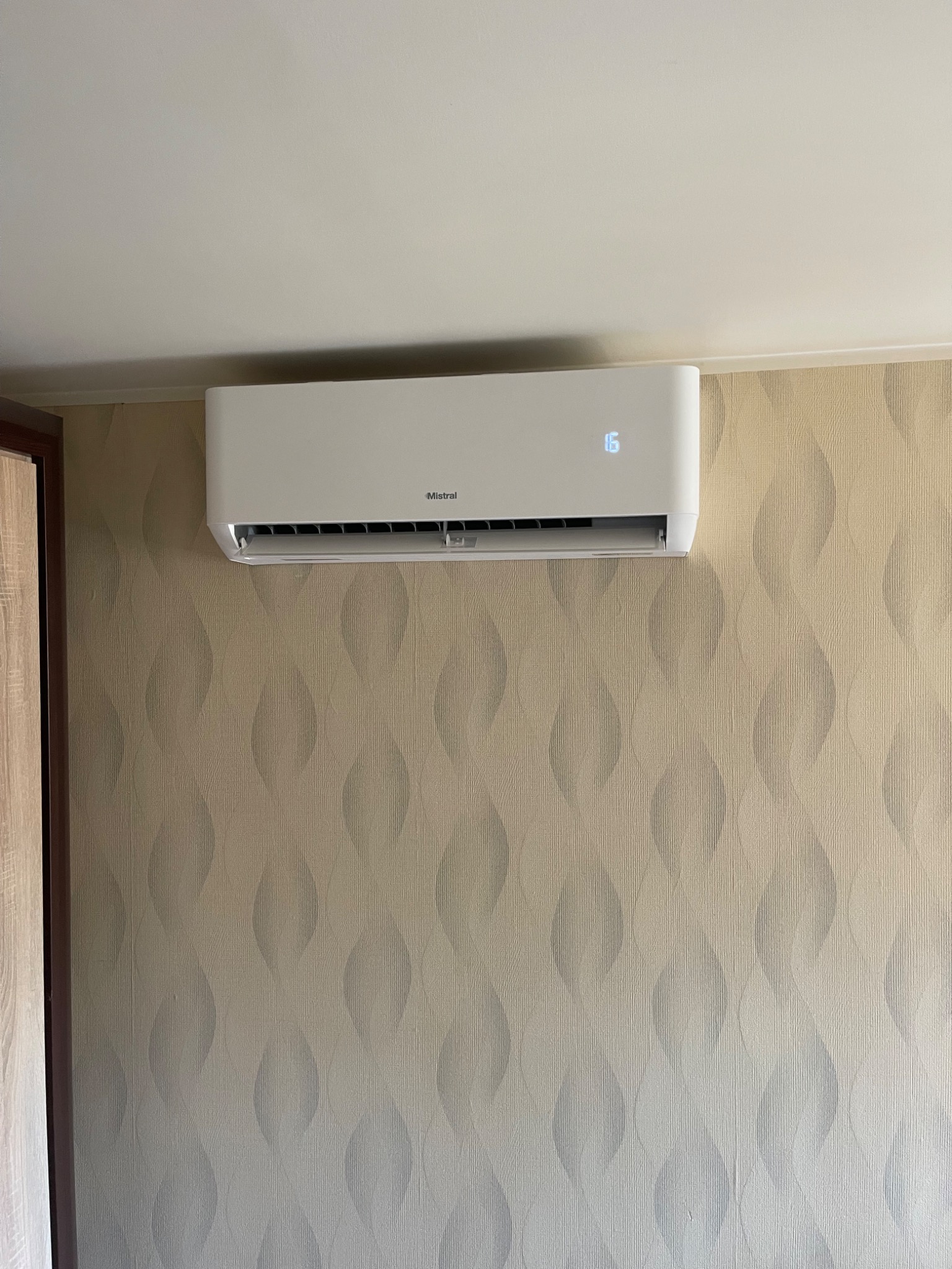 Biały klimatyzator Mistral z wyświetlaczem ustawionym na 16 stopni, zamontowany na ścianie z wzorzystą tapetą w odcieniach beżu i szarości. Widoczny fragment szafy po lewej.