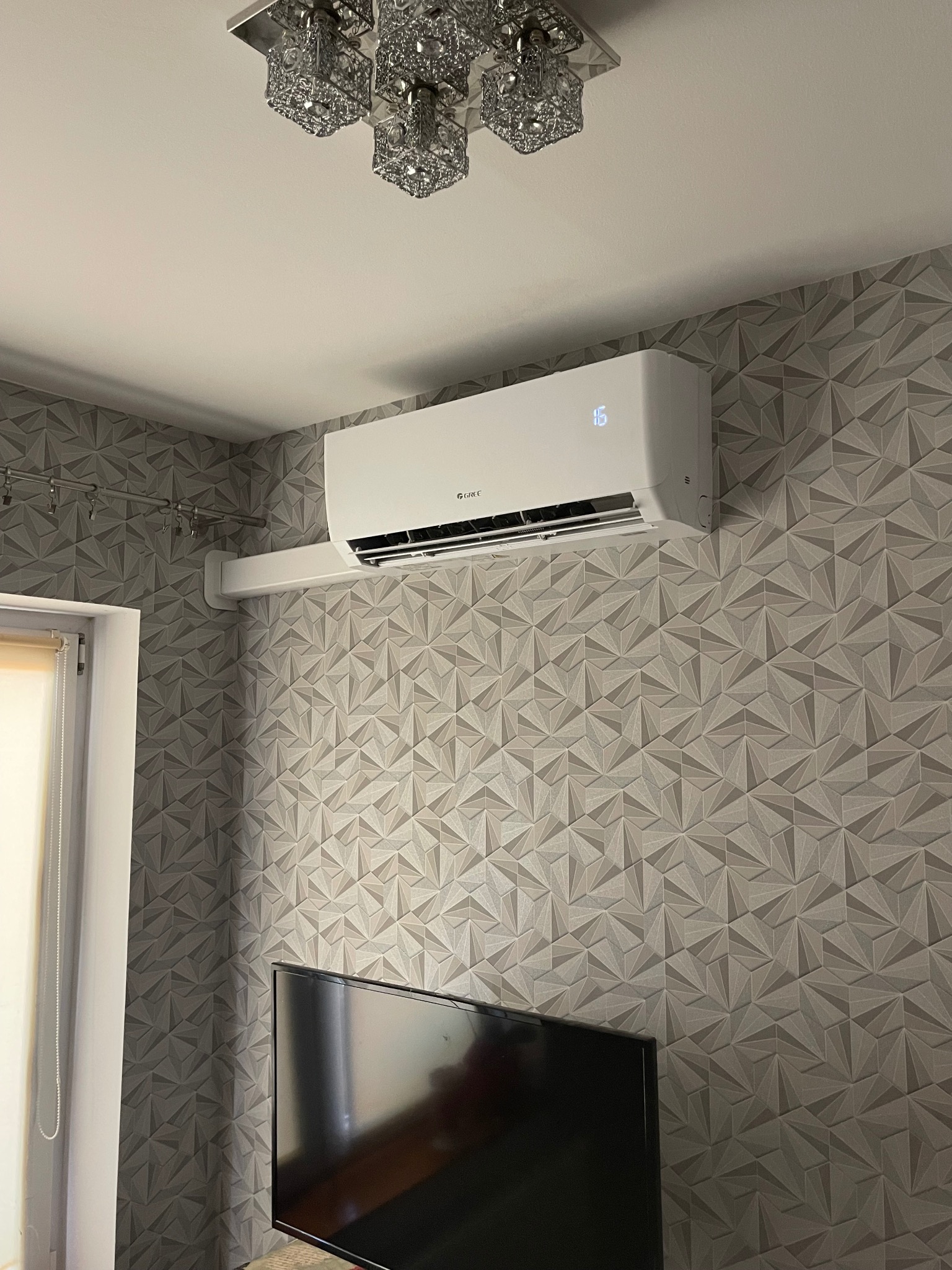 Biały klimatyzator ścienny marki Gree na ścianie z geometrycznym wzorem, pod ozdobnym żyrandolem. Widoczny fragment okna i telewizora.
