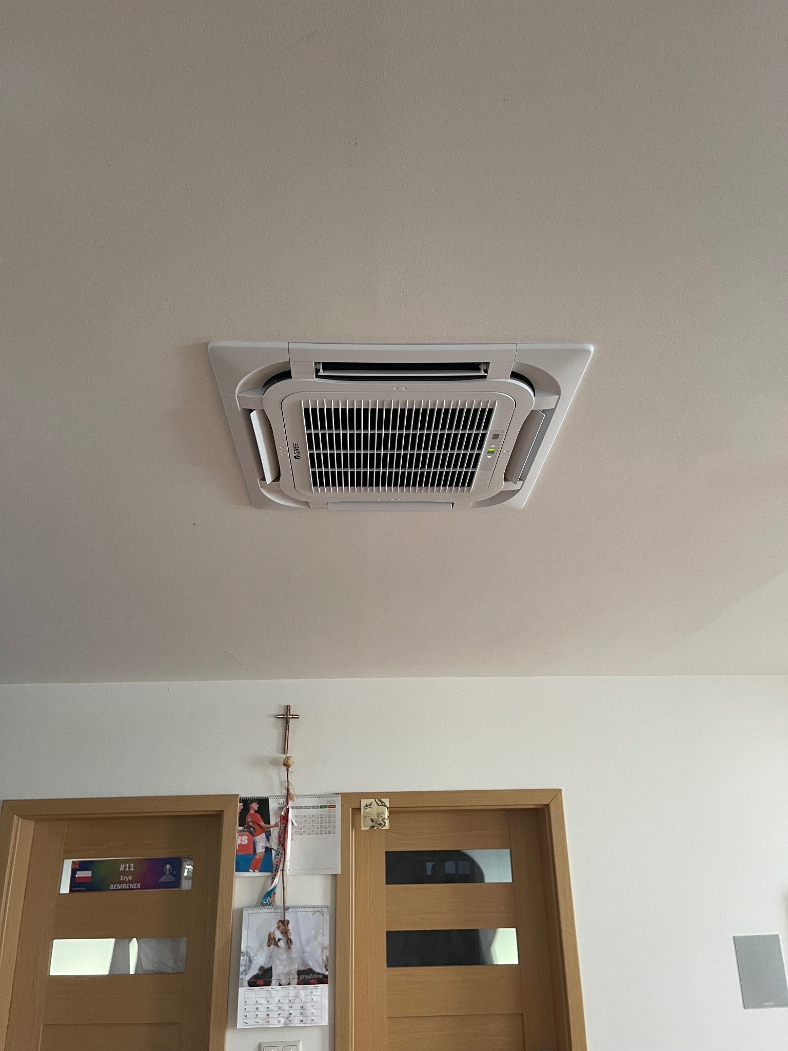 Kasetonowa klimatyzacja GREE zamontowana w suficie w Trzemesznie. Widoczne kratki wentylacyjne i biała obudowa urządzenia. Ujęcie od dołu, na ścianie krzyż i kalendarze.