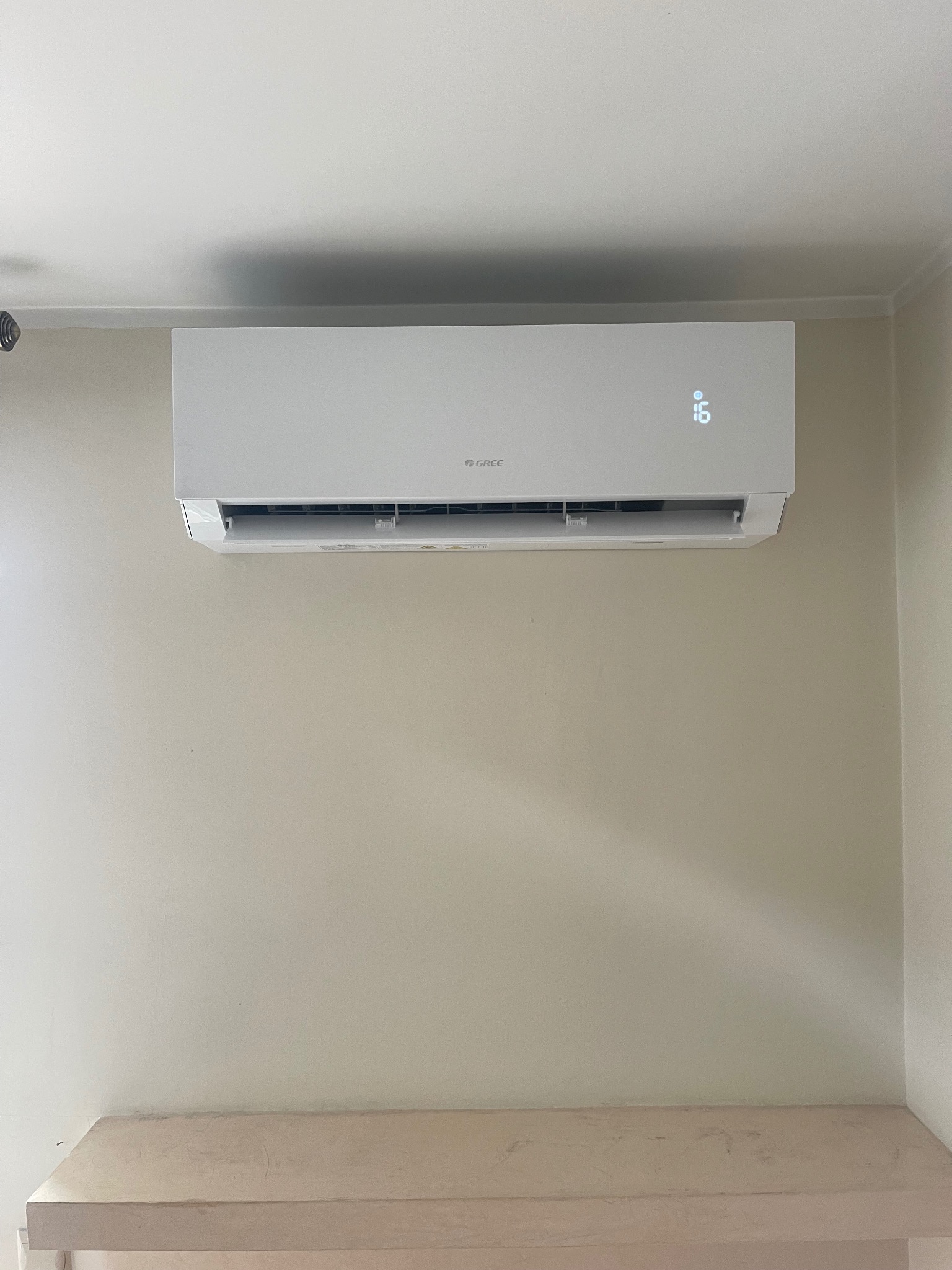 Biały klimatyzator Gree zamontowany na ścianie o kremowym odcieniu, z wyświetlaczem pokazującym temperaturę 16 stopni. Na dole widoczny fragment jasnego blatu.