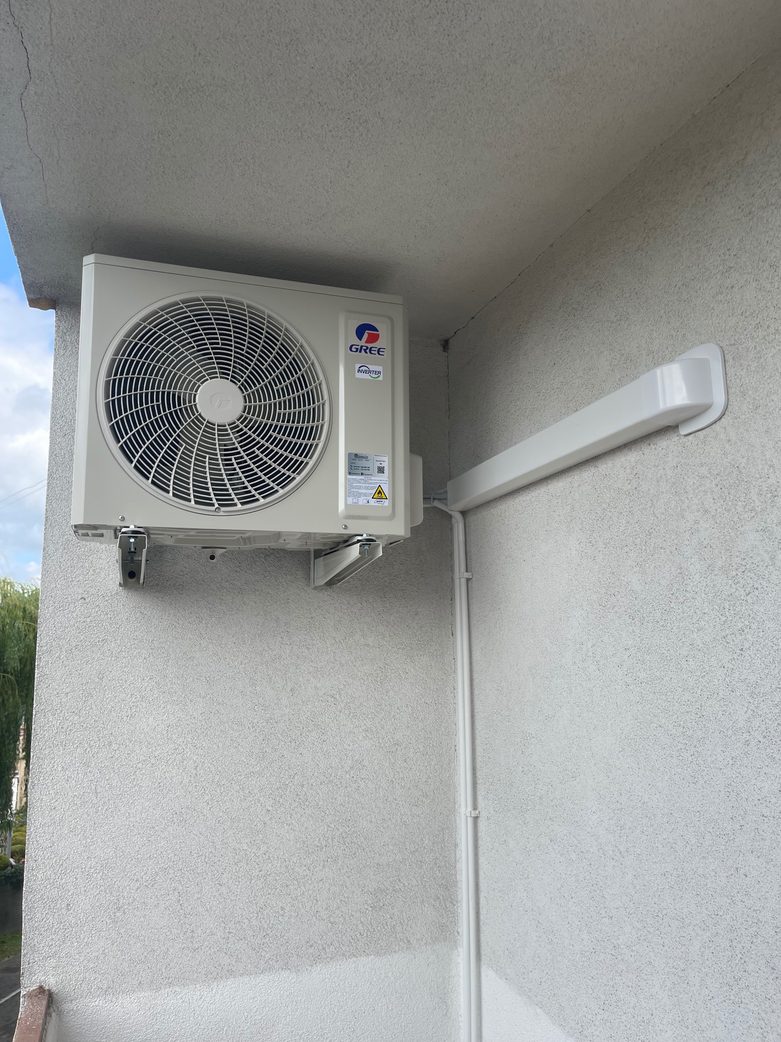 Zamontowana jednostka zewnętrzna klimatyzatora Gree na elewacji budynku z widocznymi przewodami w białej osłonie. Widok z dołu, szara elewacja.