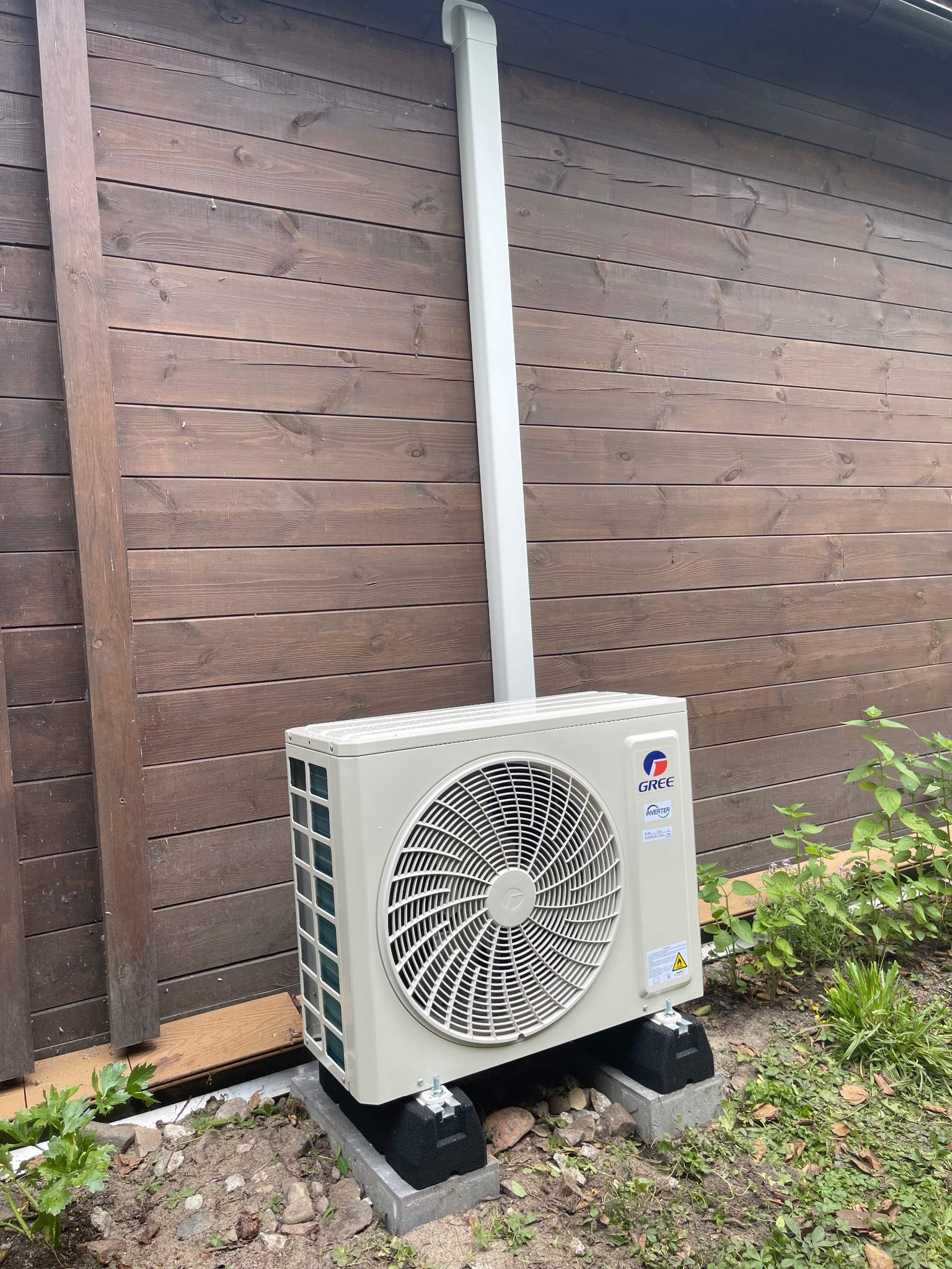 Zamontowana jednostka zewnętrzna klimatyzacji Gree na betonowych podstawach przy drewnianej ścianie budynku z rurą odprowadzającą. Widoczne rośliny w tle.
