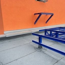 EKOOBUDOWA SPÓŁKA Z OGRANICZONĄ ODPOWIEDZIALNOŚCIĄ - Szare pokrycie dachowe z widoczną konstrukcją wsporczą w kolorze niebieskim na tle pomarańczowej ściany budynku. Widok z góry, oświetlenie dzienne.