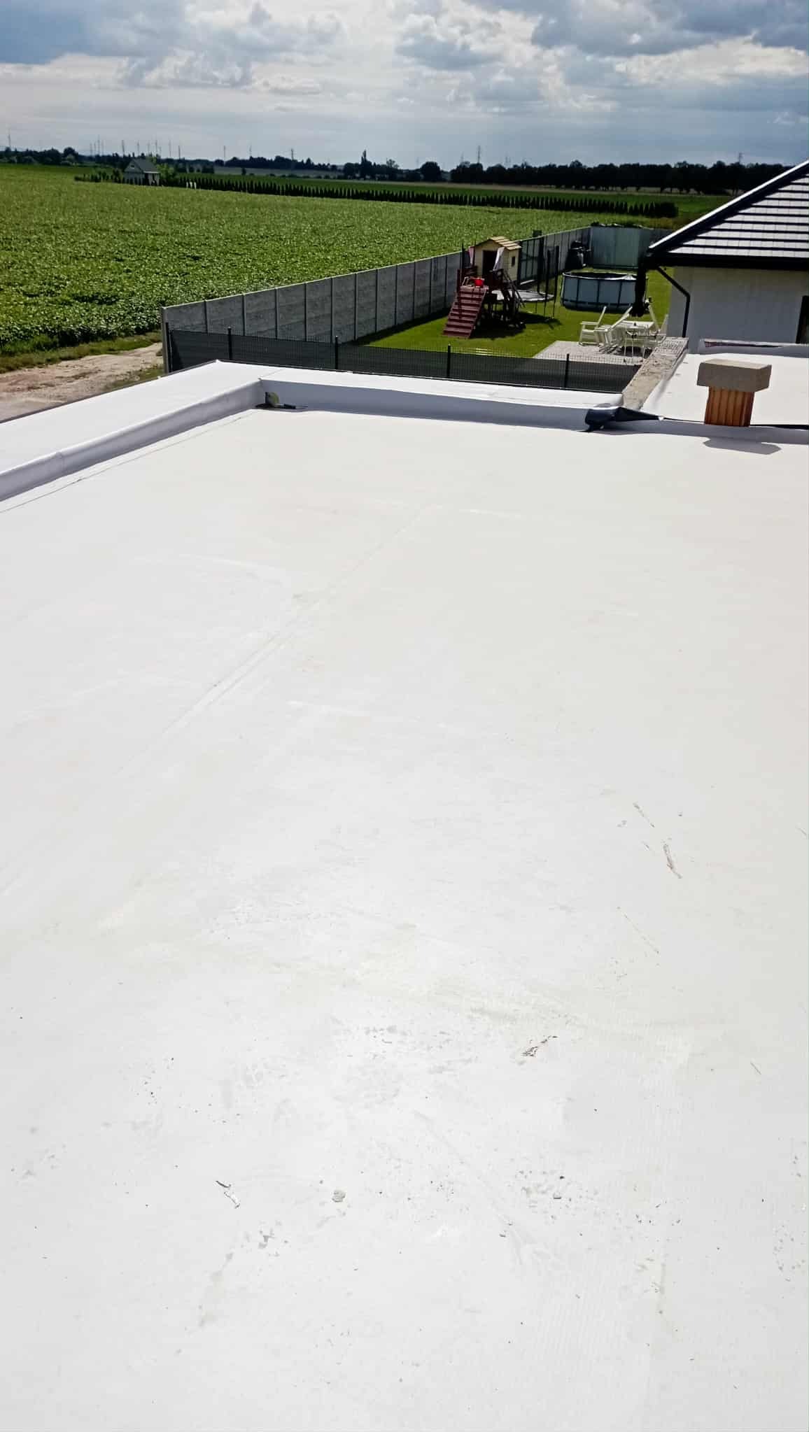 Biały, płaski dach z wykończonymi krawędziami, widok z góry na tle zielonego pola i zabudowań. Nowoczesne pokrycie dachowe, detale wykończeniowe.