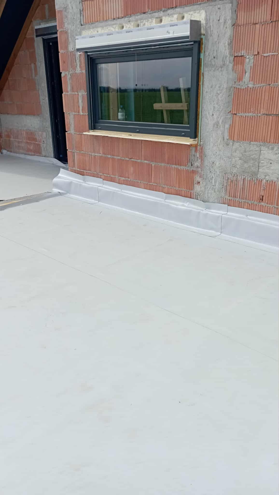 Wykończony dach płaski z obróbkami blacharskimi przy ścianie z pustaków ceramicznych i oknem. Widoczna membrana dachowa w jasnym kolorze.