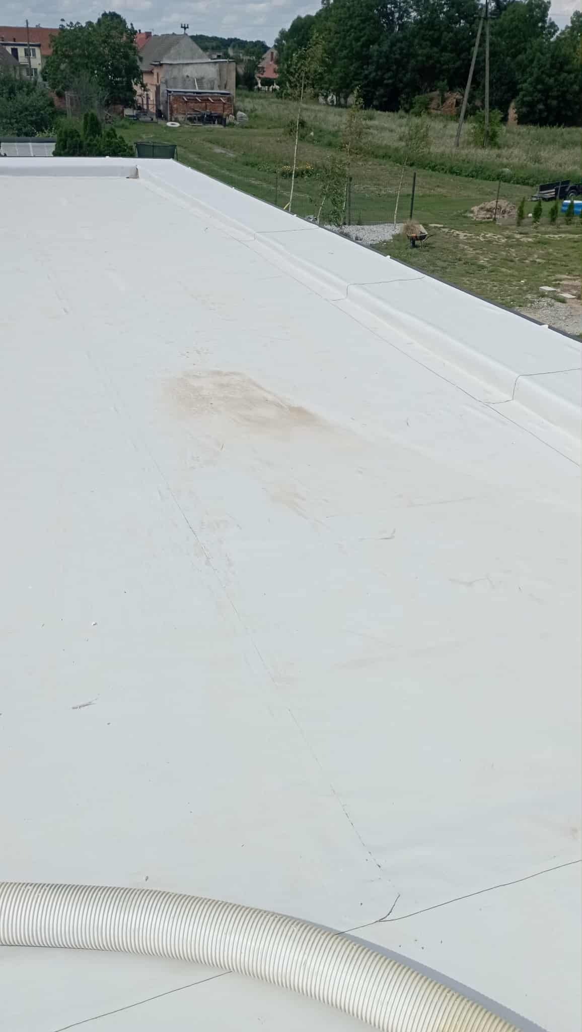 Biały, płaski dach budynku po hydroizolacji z widocznym przewodem wentylacyjnym i fragmentem otoczenia w tle.