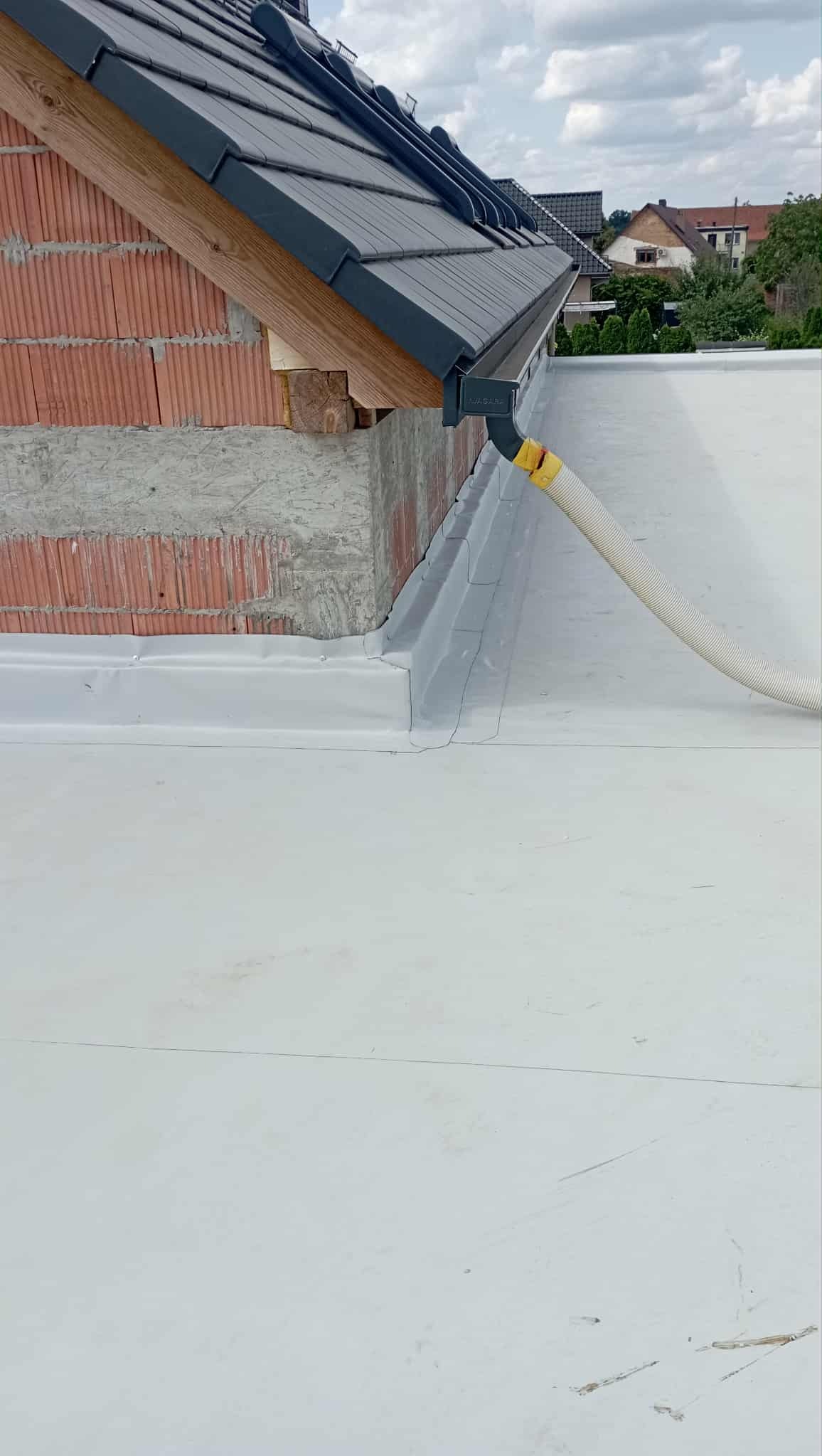 Szary dach z rynną i białą membraną dachową na surowej cegle. W tle domy i drzewa.