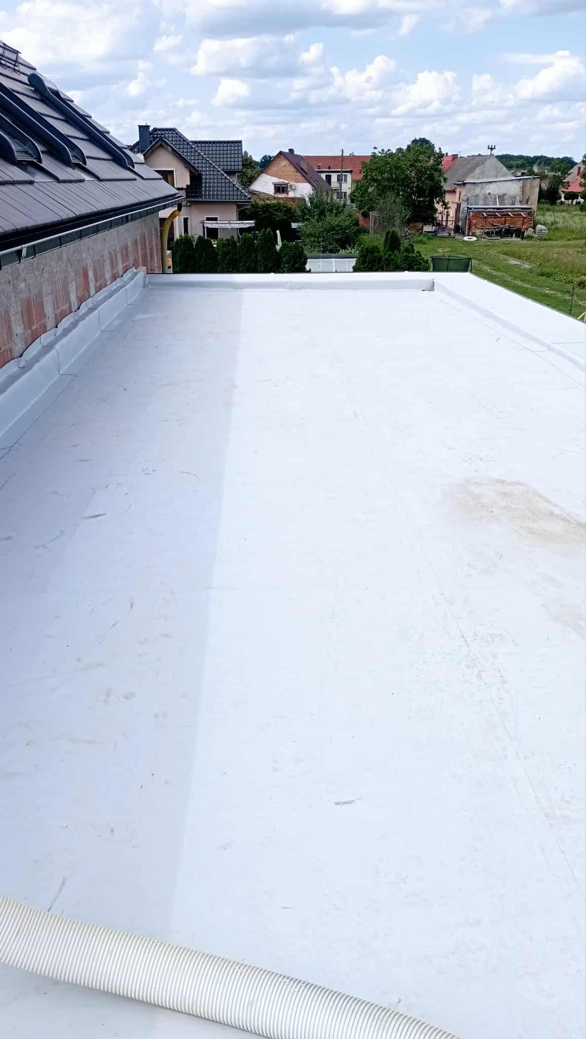 Biała, płaska powierzchnia dachu po kryciu, z widocznym fragmentem dachu skośnego, murem i zabudową w tle. Widoczny element wentylacji. Słoneczny dzień.